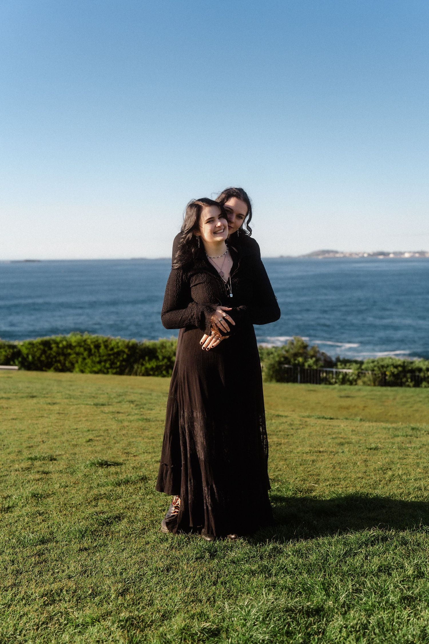A couple embraces on a grassy oceanside cliff with blue water in the background.