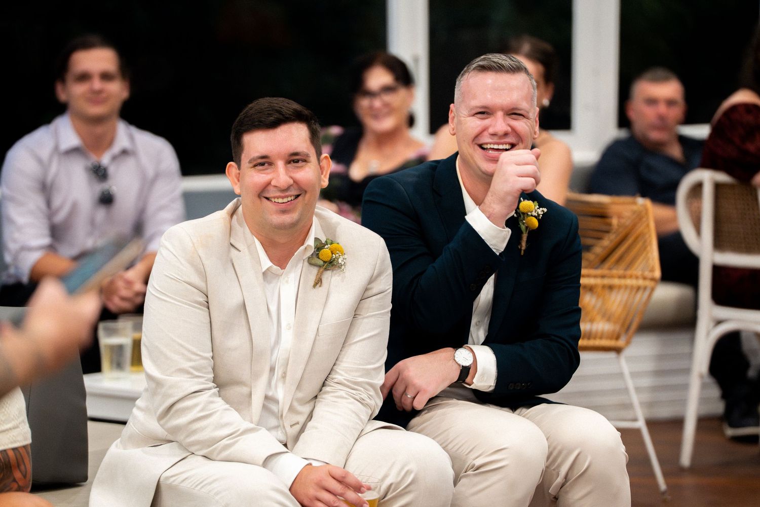 Two grooms smiling joyfully at their wedding ceremony, one in cream suit and one in navy blazer with boutonniere.