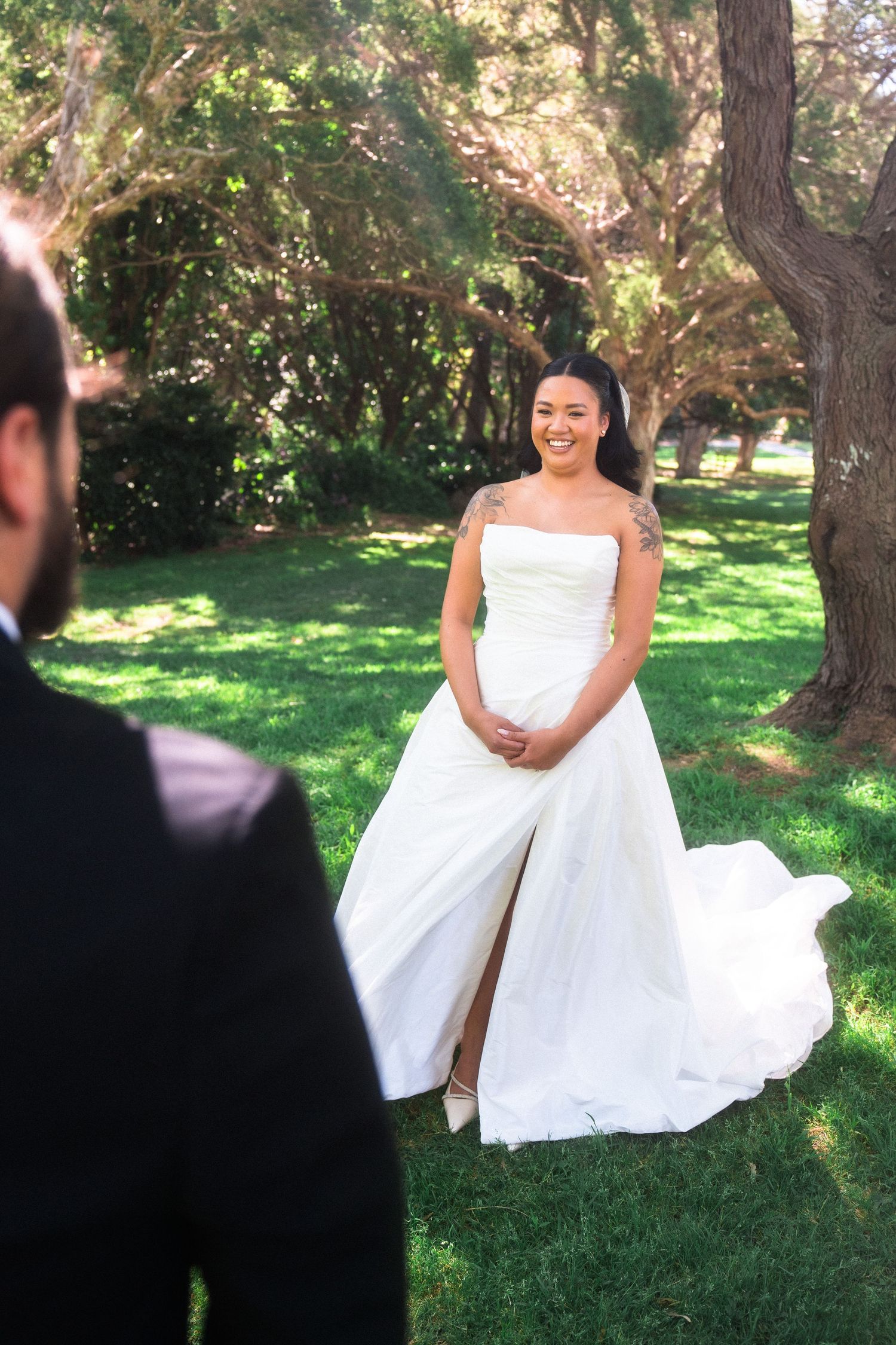 A bride in a white dress poses on grass under shaded trees.