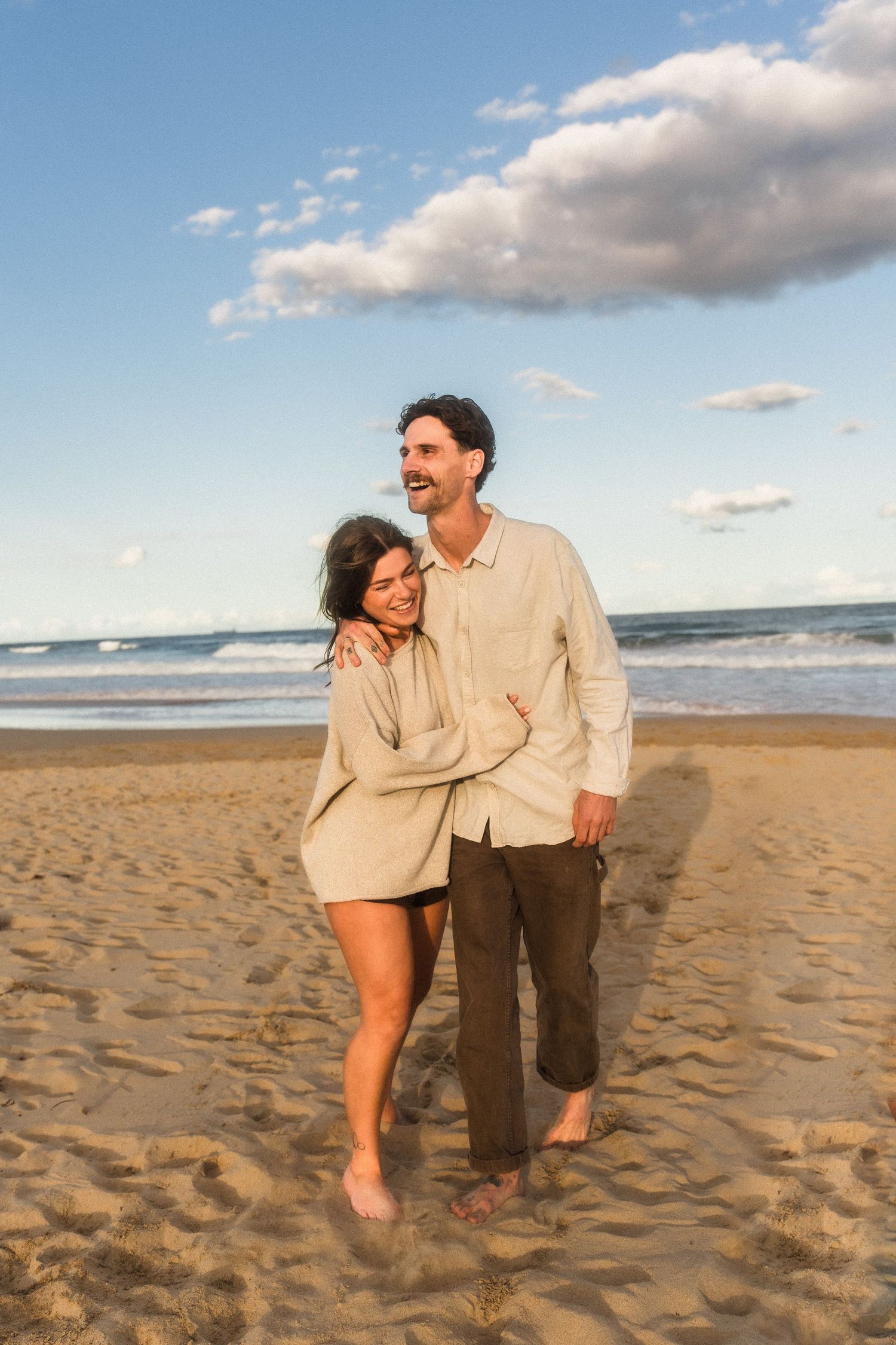 Couple walking and laughing together on a sandy beach during sunset.