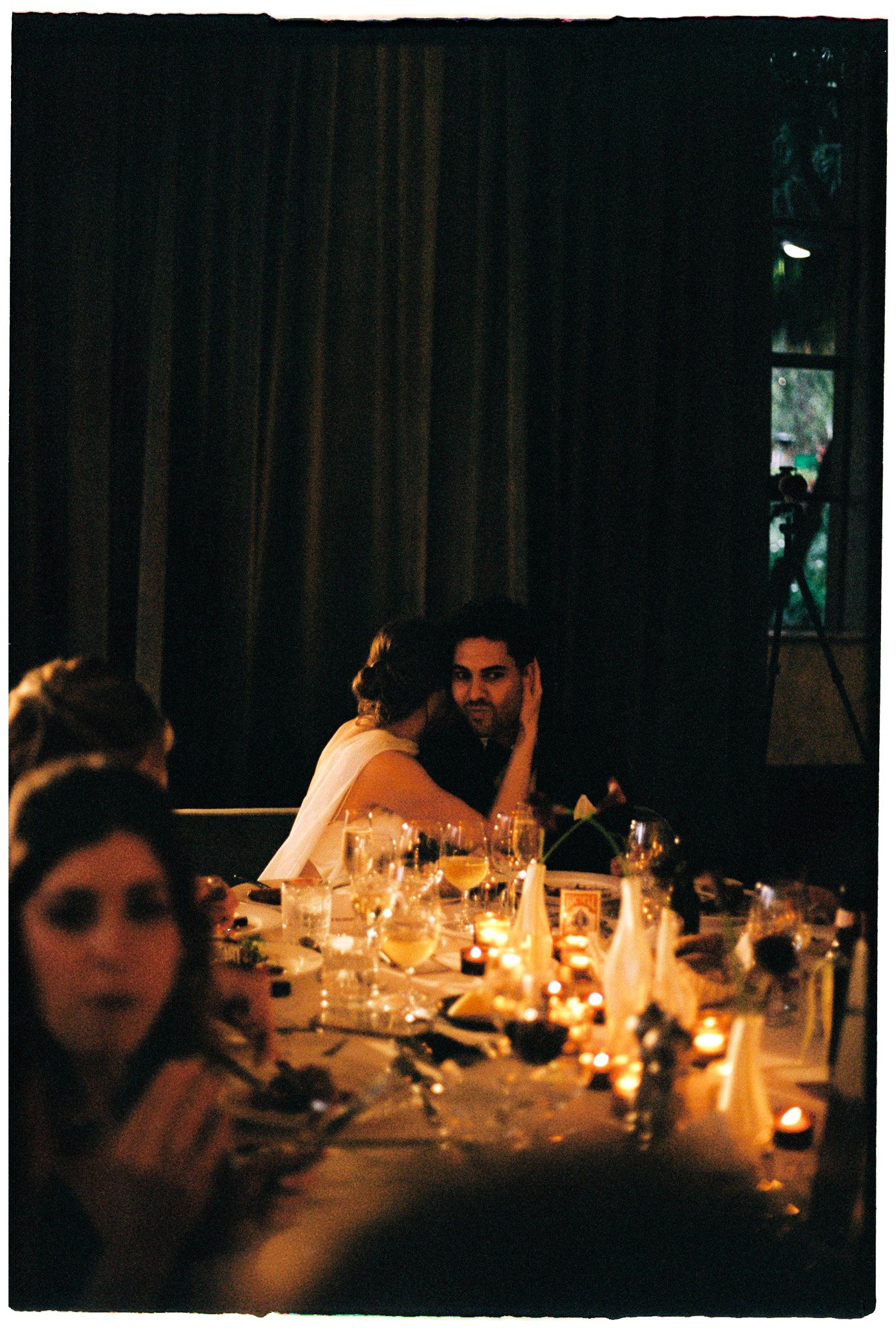 Candlelit wedding reception dinner table with guests in dim romantic lighting, featuring elegant table settings and glowing candles.