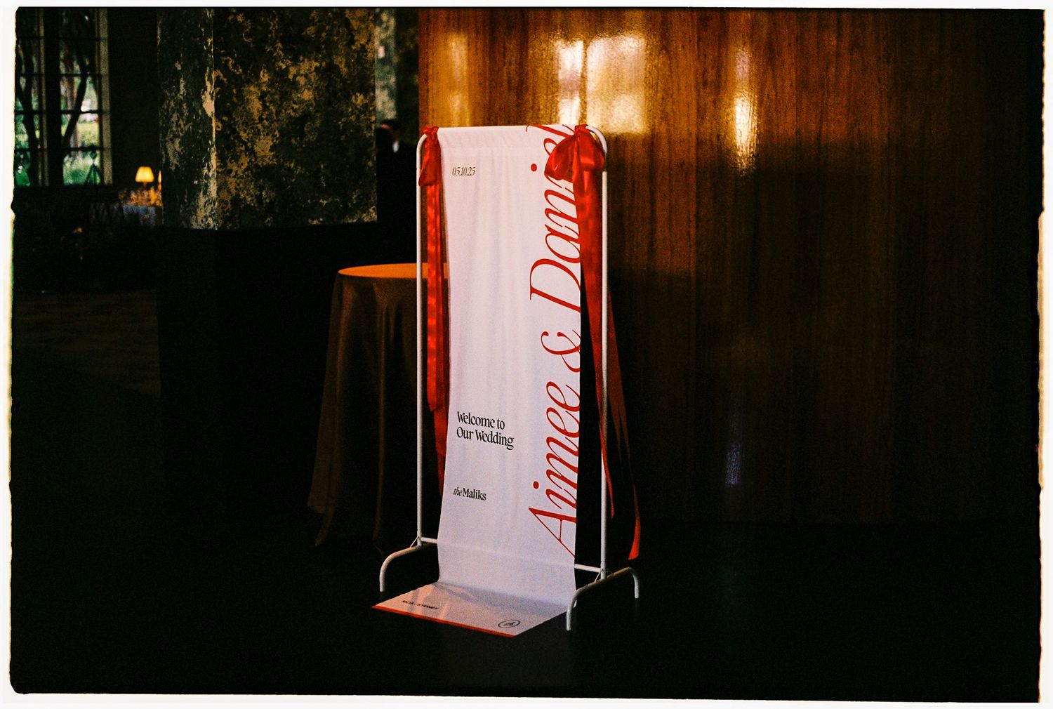 Elegant wedding signage banner displaying Aimee & Dan on a metal stand in a dimly lit reception venue.