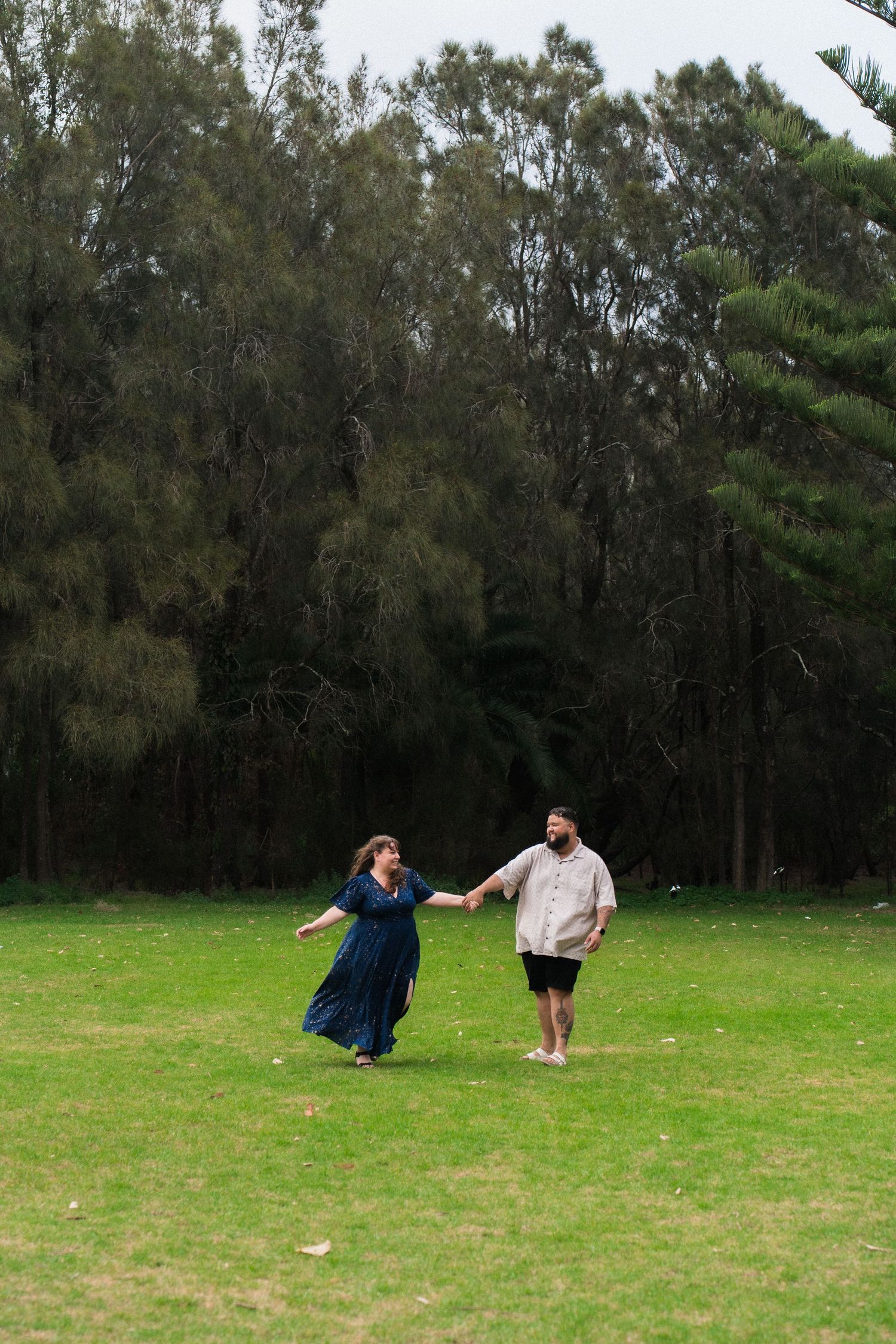 Playful couple holding hands while spinning in circles on a grassy field surrounded by trees.