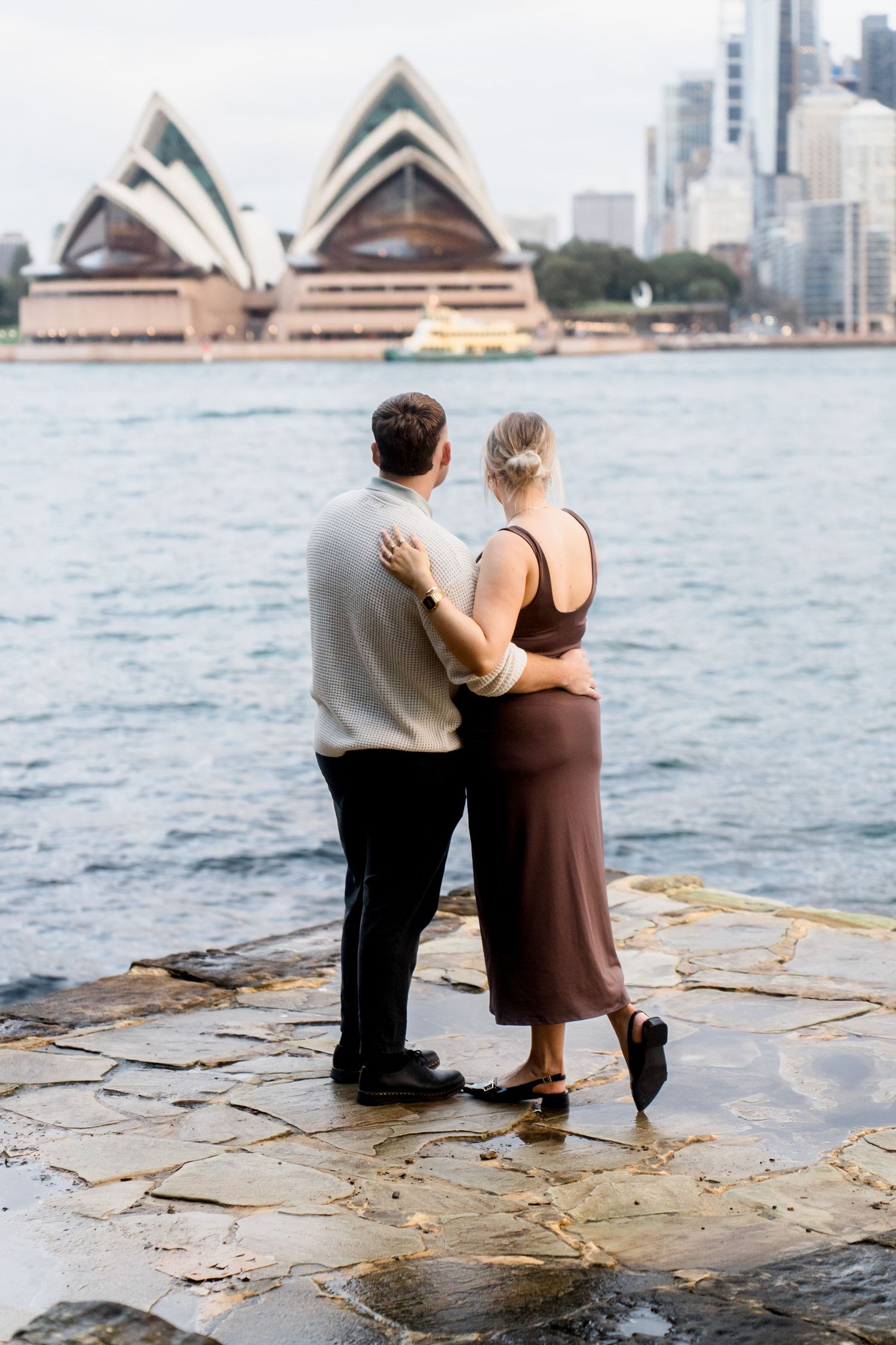 A couple admires the Sydney Opera House while standing on rocky shoreline.