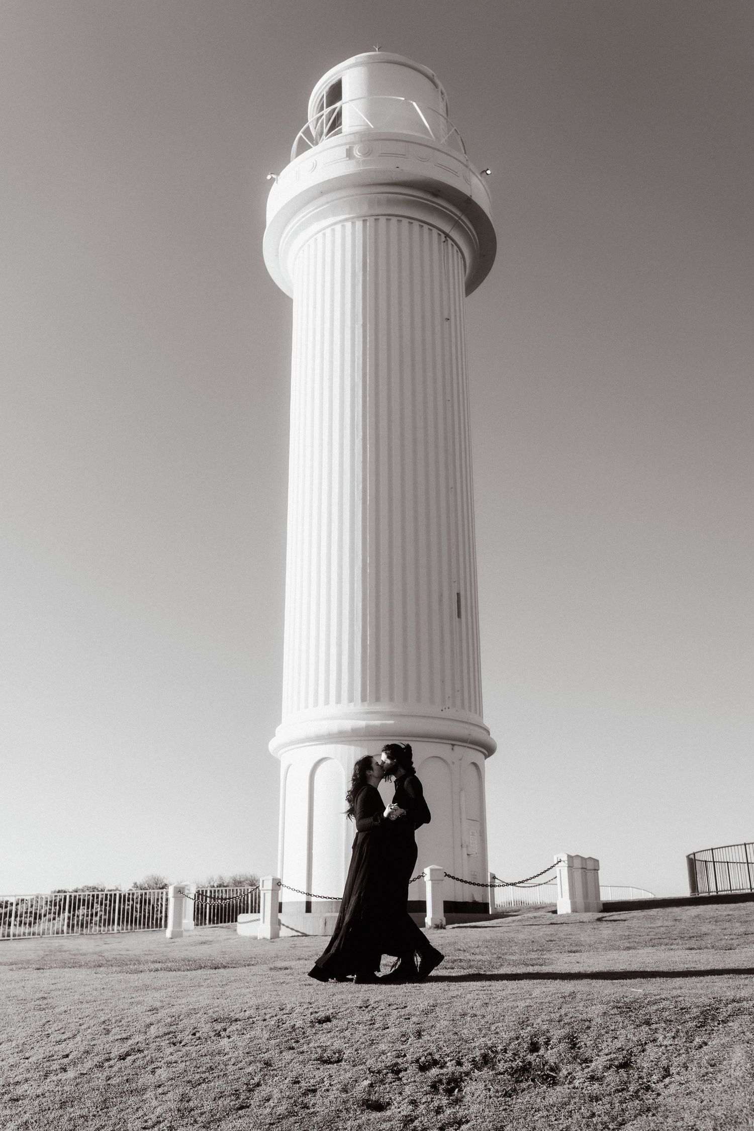Two silhouettes stand near a white lighthouse tower against a minimalist backdrop.