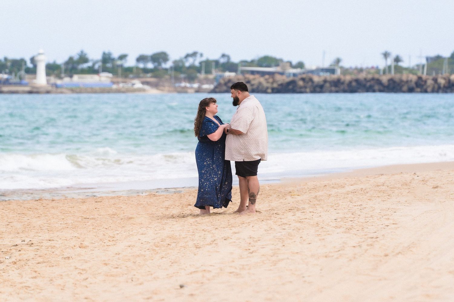 Couple sitting together on a sandy beach with blue ocean waters and coastal scenery.