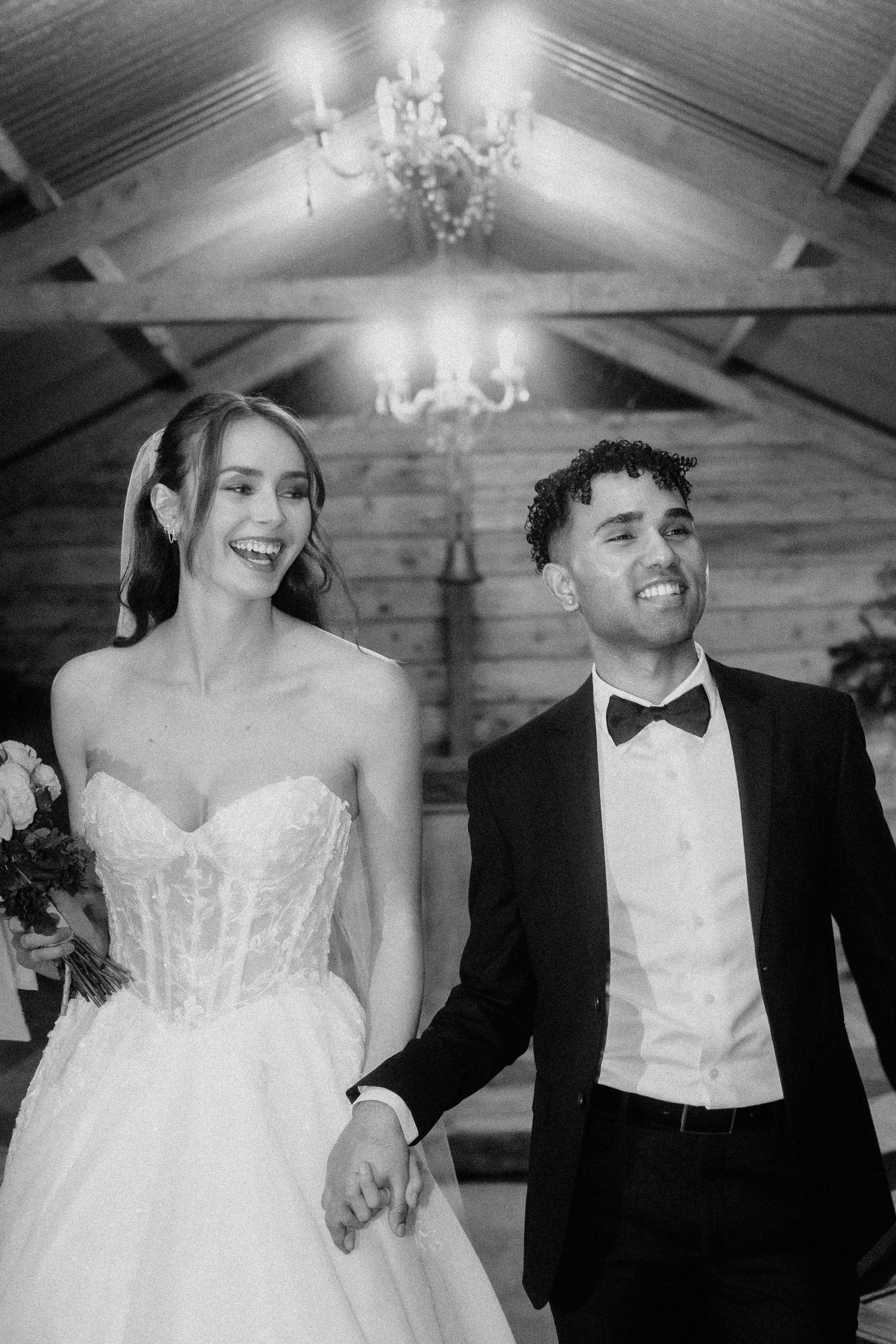 A joyful wedding couple dances together under a wooden beam ceiling with elegant chandeliers.