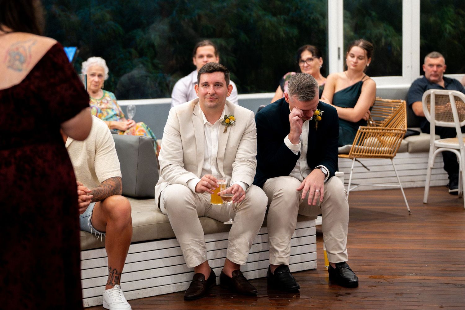 Two grooms sit together during wedding ceremony, one emotional, guests seated behind them outdoors.
