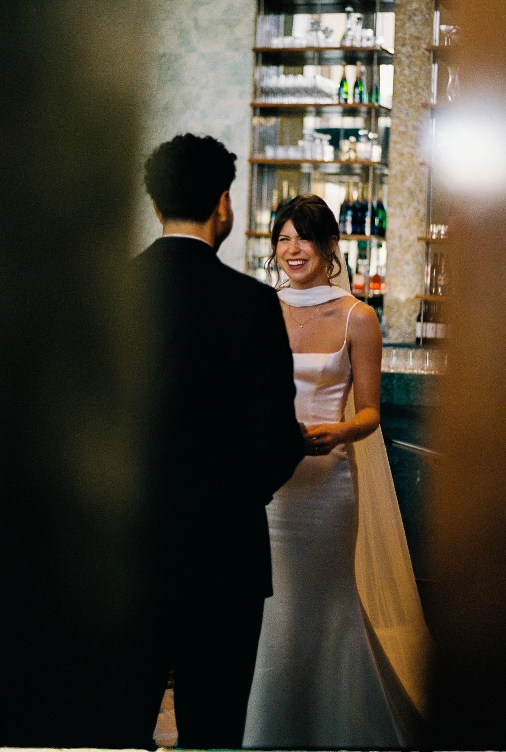 Bride in elegant white gown smiling at groom in black suit near a bar, capturing a romantic wedding moment.