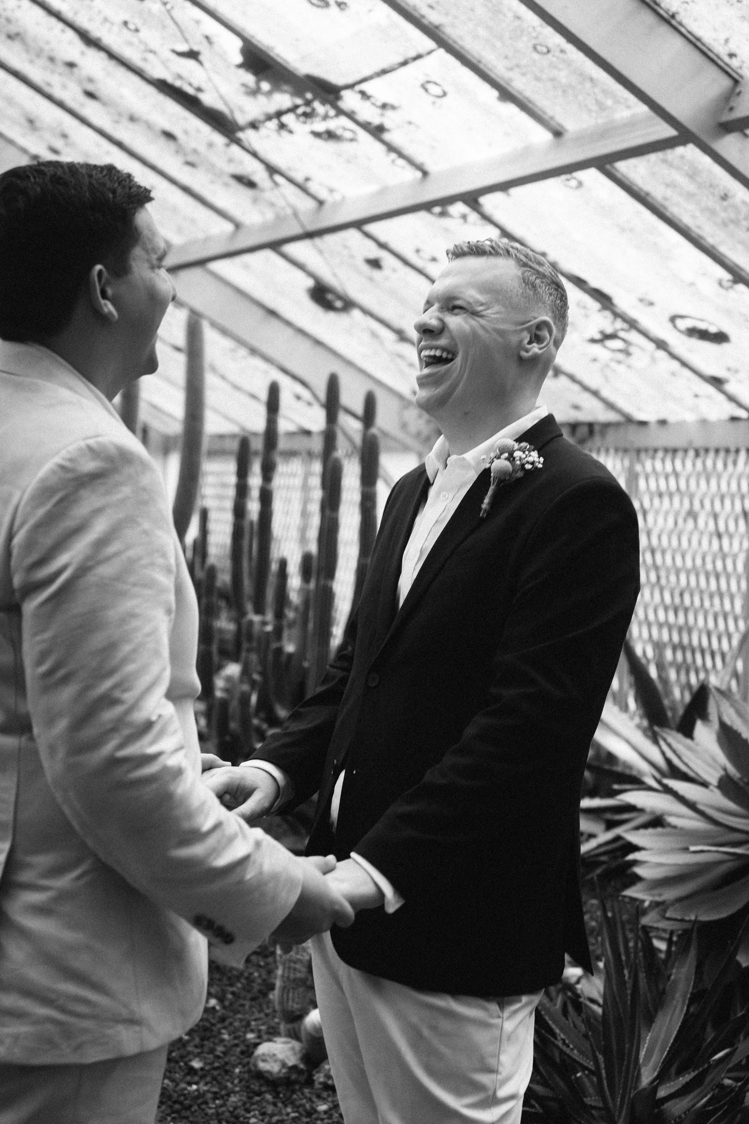 Two grooms laughing and holding hands in a greenhouse with cacti, black and white wedding photo.