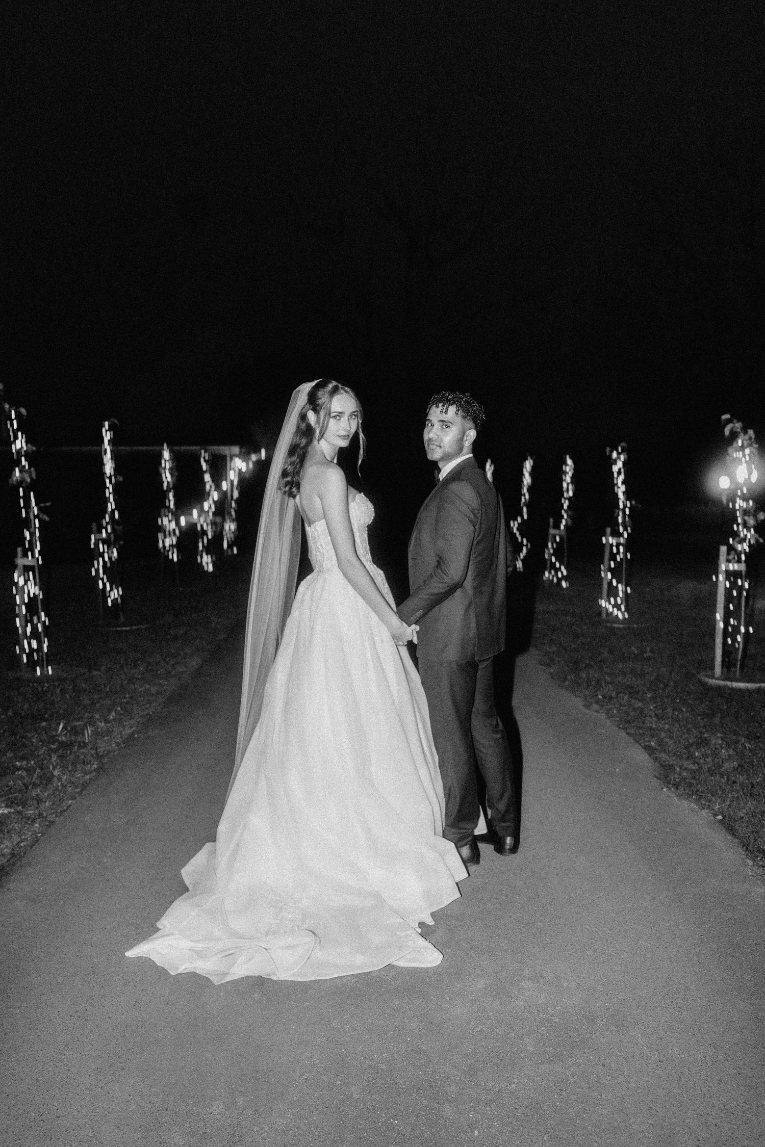 A wedding couple poses in an outdoor evening setting with string lights in the background.