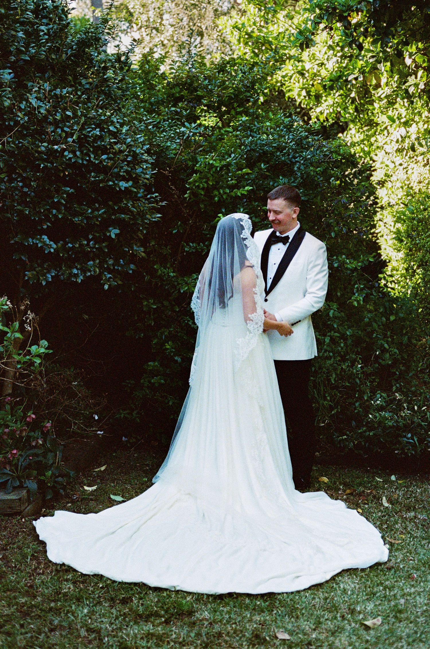 Wedding couple poses in a garden setting with flowing white dress and classic tuxedo.