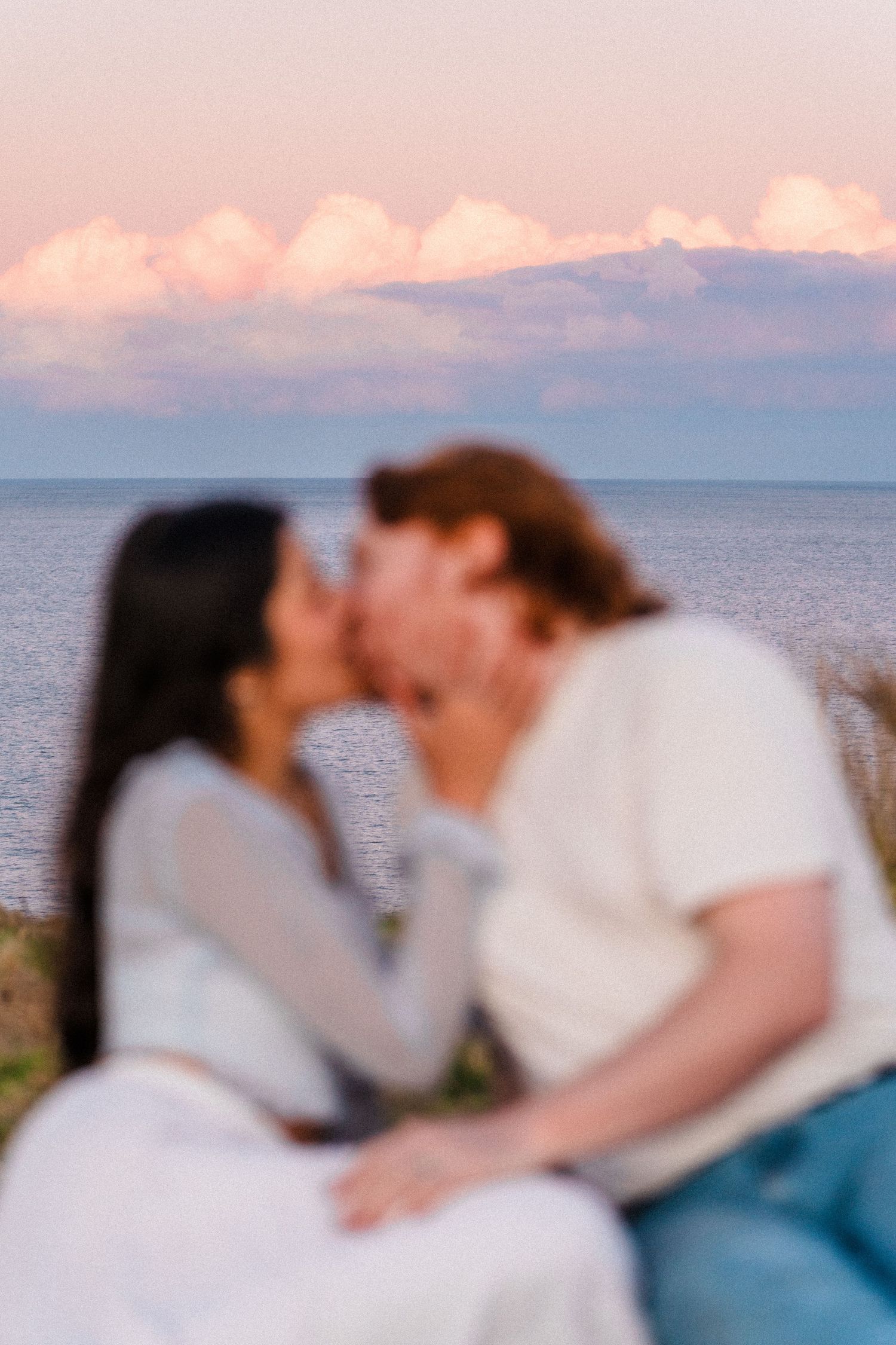 A romantic couple shares a tender kiss against a sunset ocean backdrop with pink clouds.