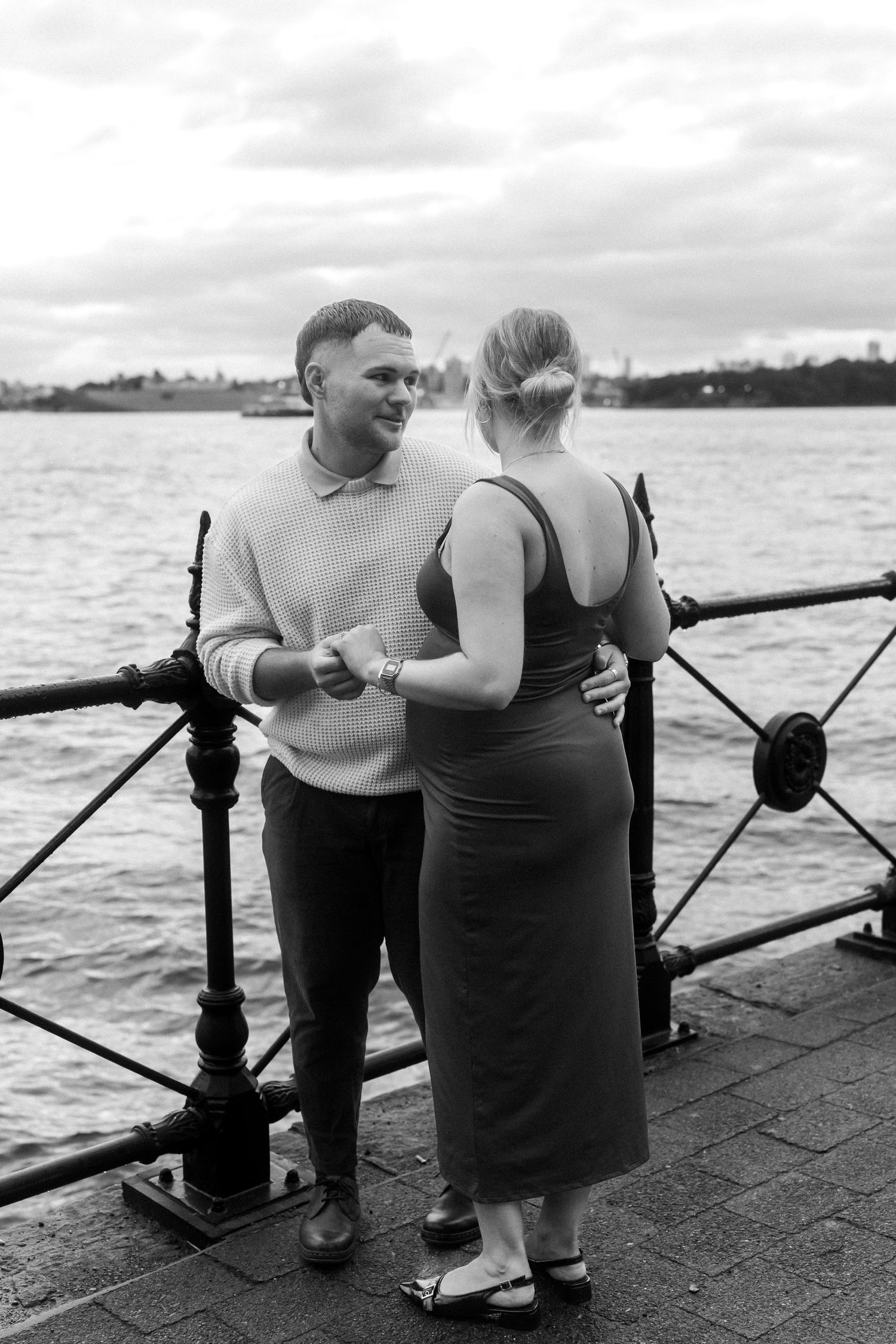 A couple shares an intimate moment along a waterfront railing in black and white.