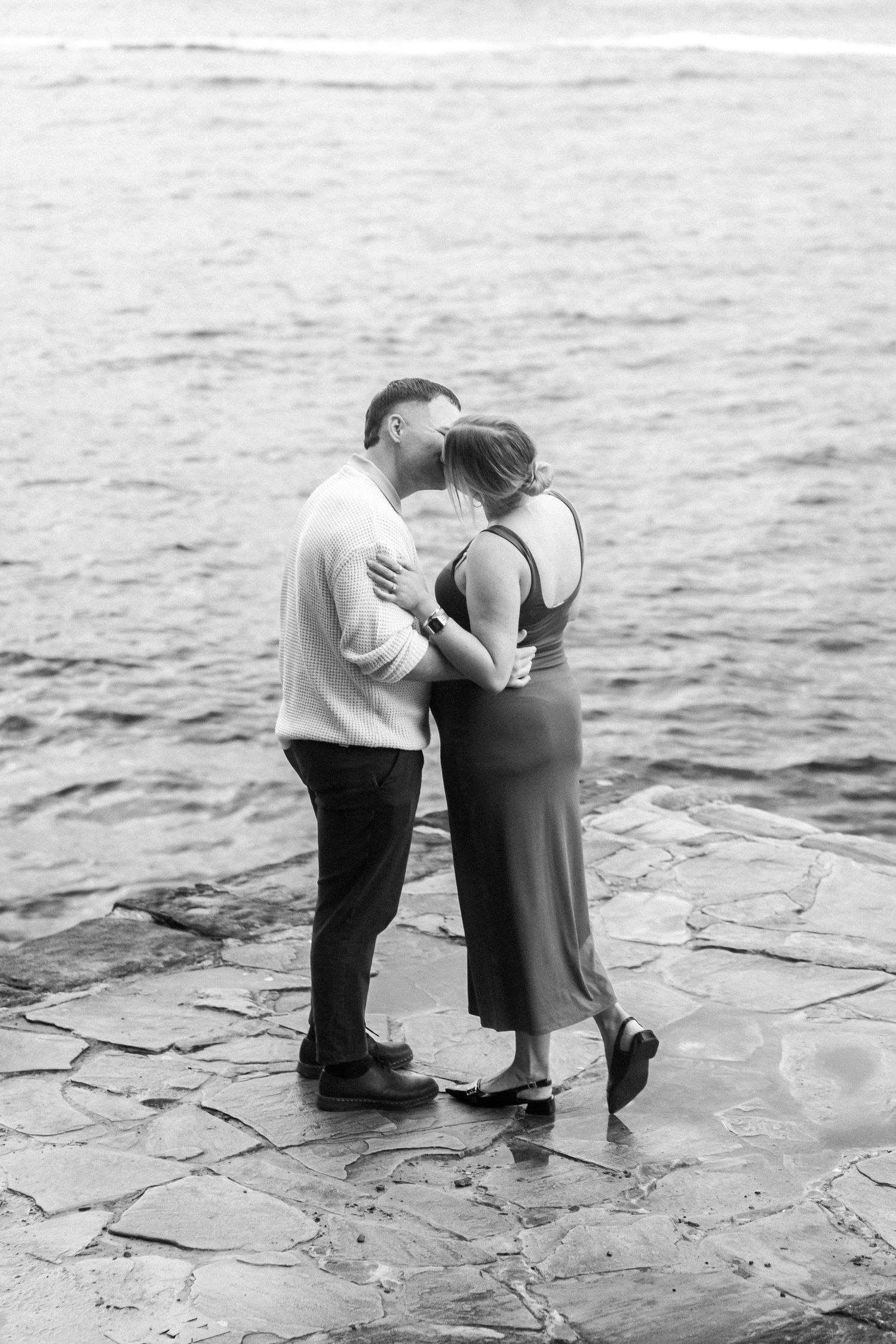 A black and white photo of a couple embracing on stone waterfront.