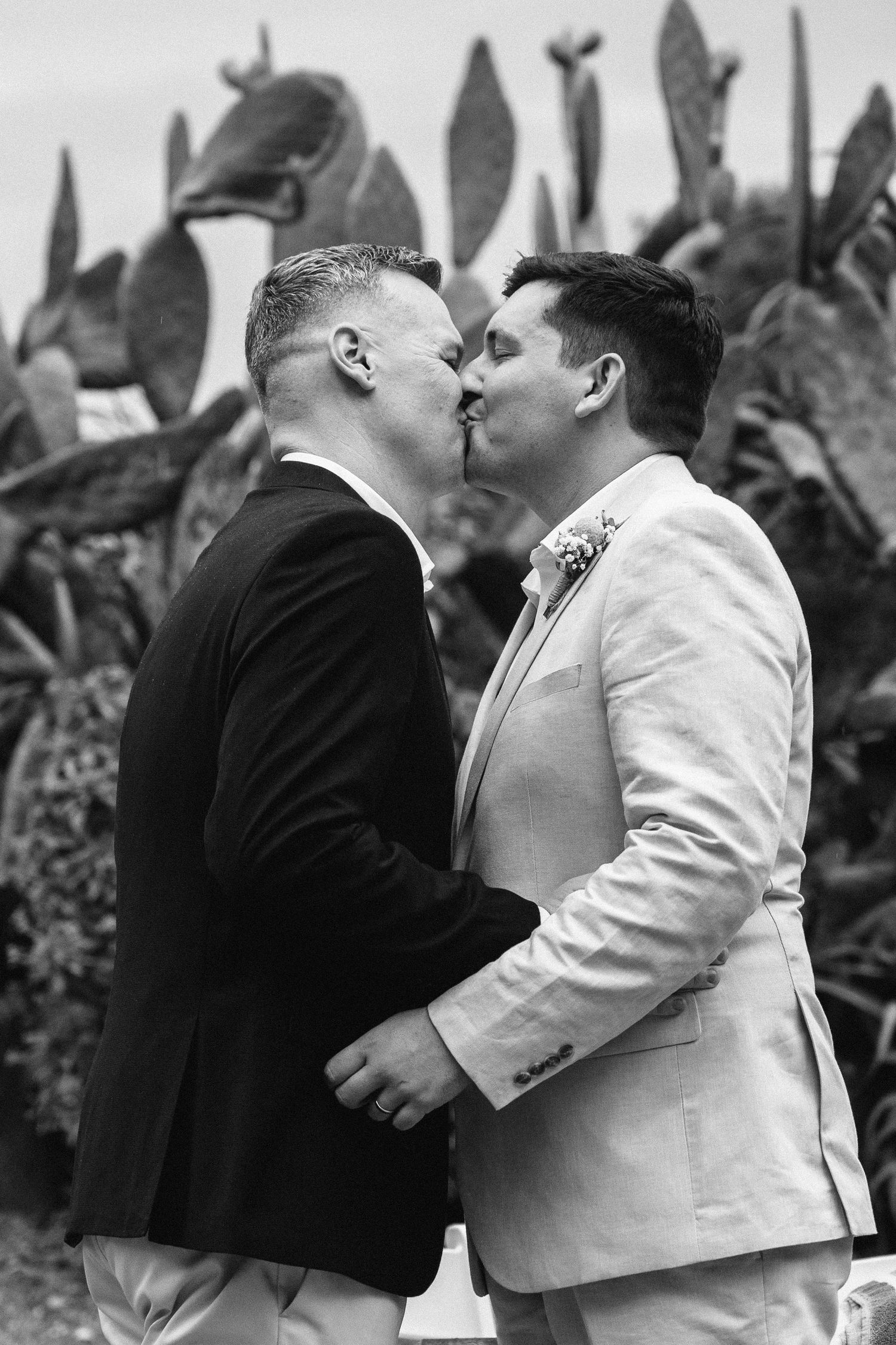 Black and white photo of two grooms sharing a kiss at their wedding ceremony with cactus backdrop.