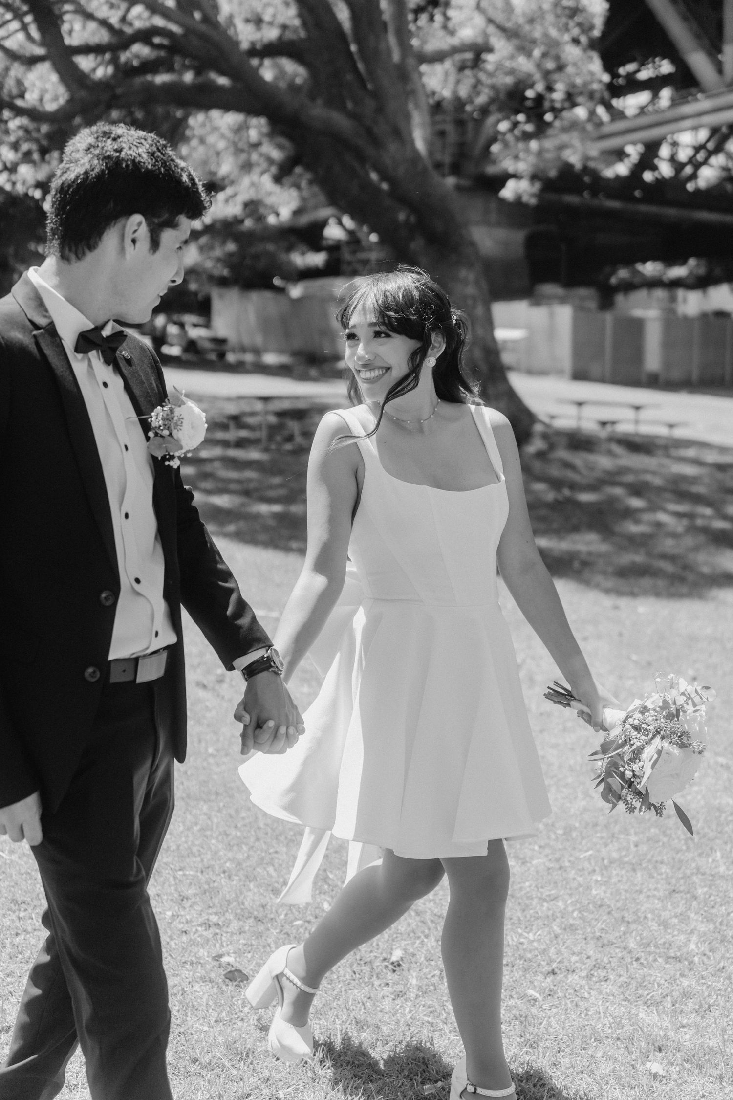 A playful couple walks together on grass in black and white wedding photography.