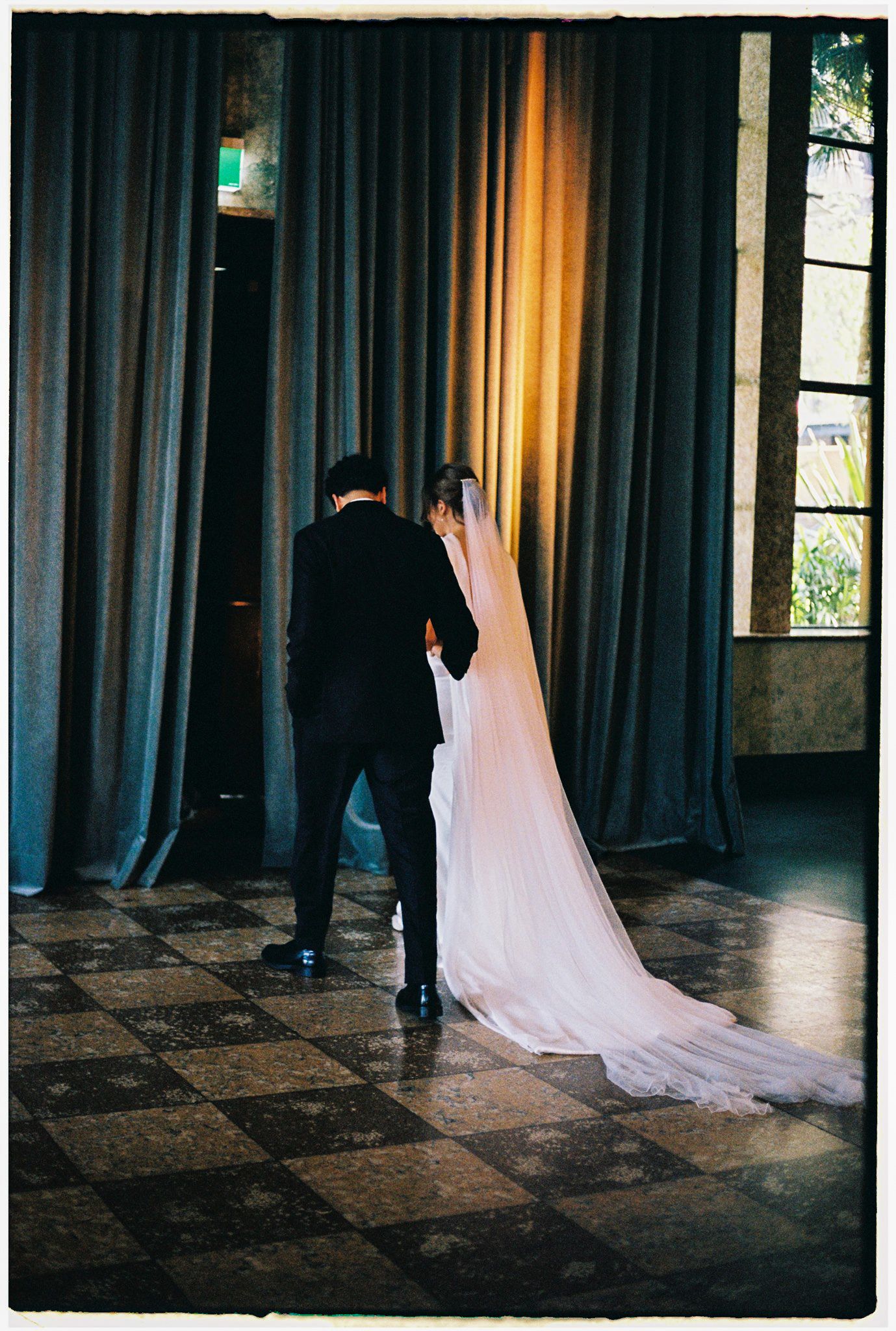 Bride and groom share an intimate moment, her long veil trailing on a checkered floor near draped curtains.