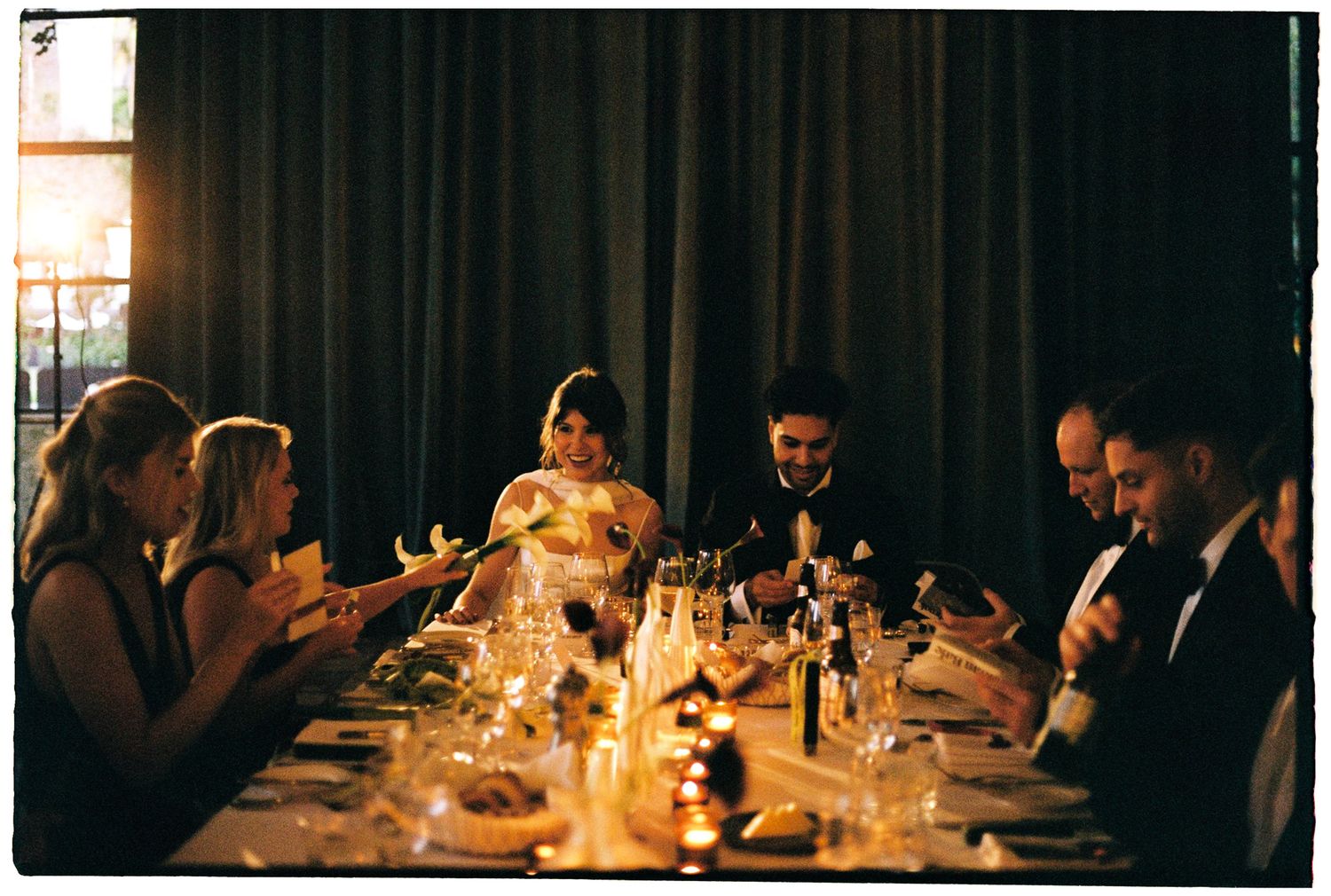 Elegant candlelit dinner party with guests in formal attire seated at a long table with wine glasses and soft lighting.