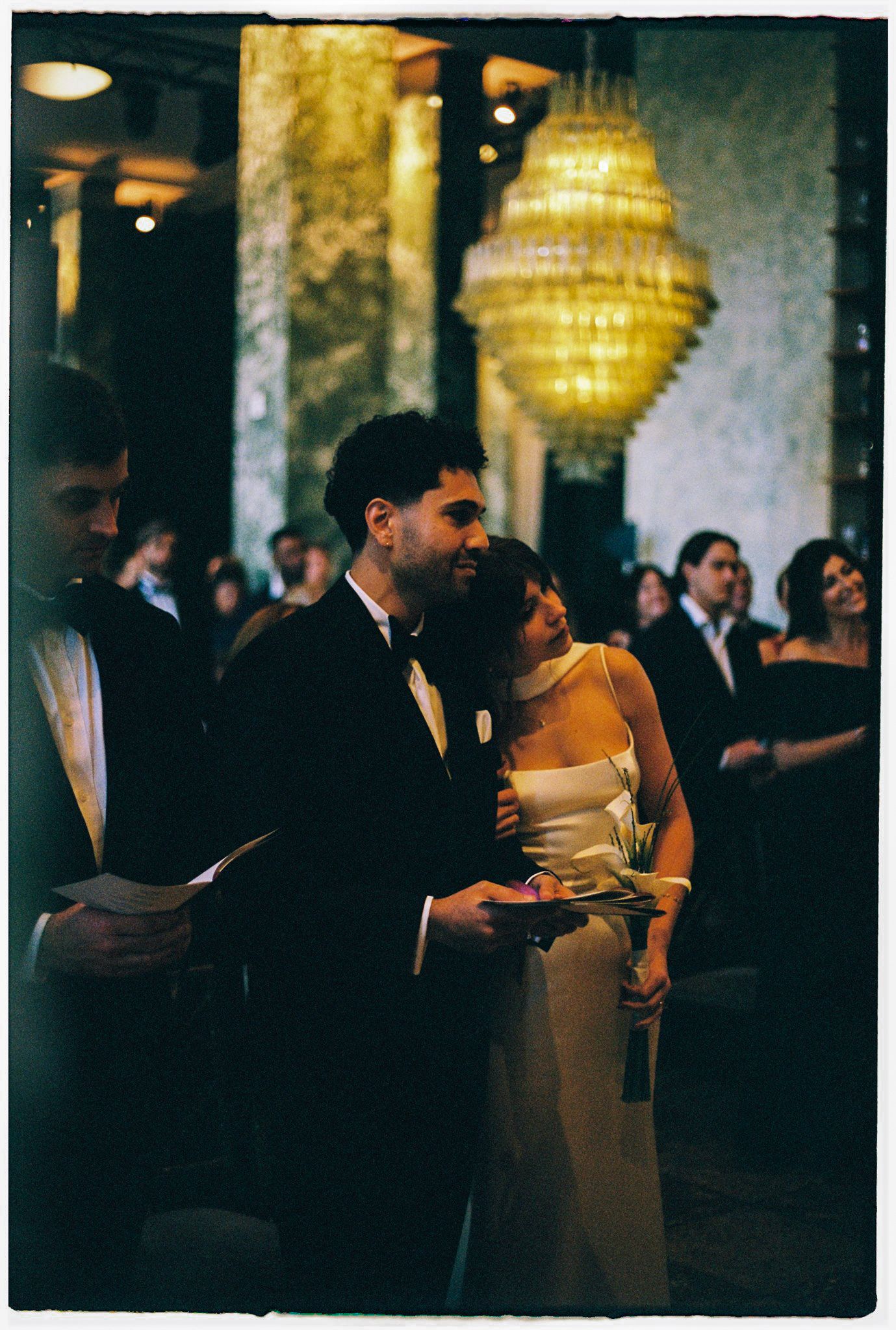 Bride and groom share a tender moment during their wedding ceremony, with golden chandeliers glowing above.