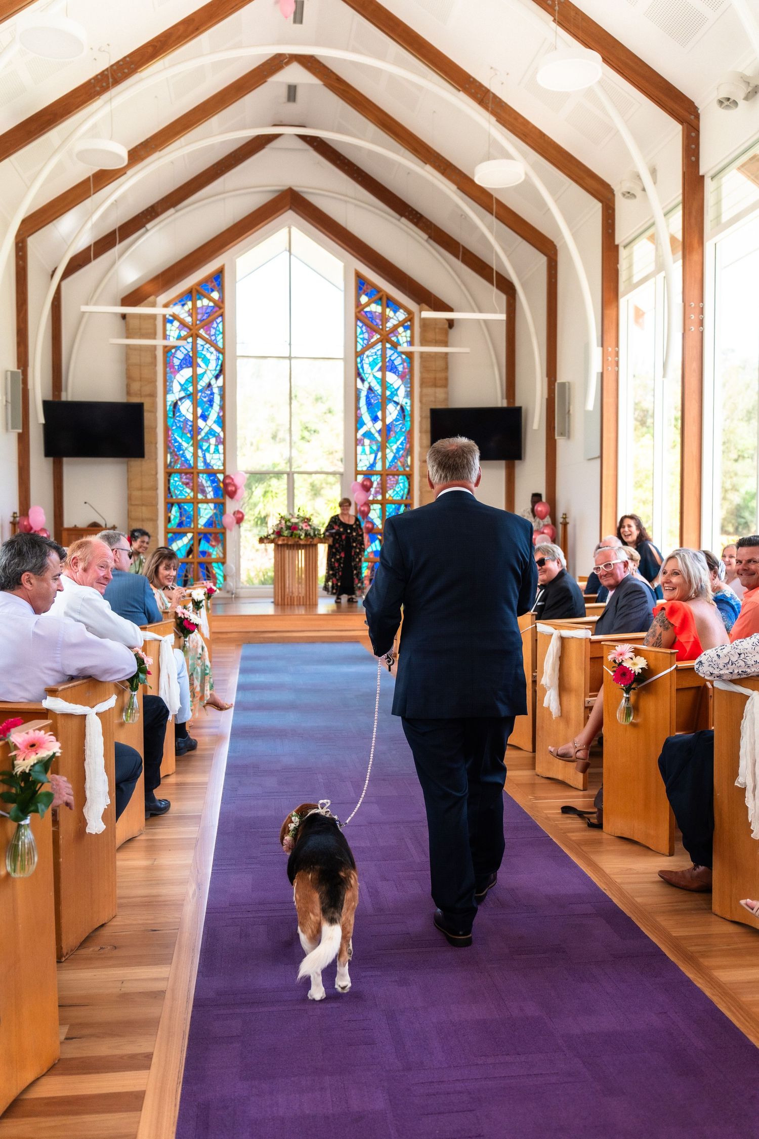 Wedding ceremony in a church with wooden beams and purple carpet leading to stained glass windows.