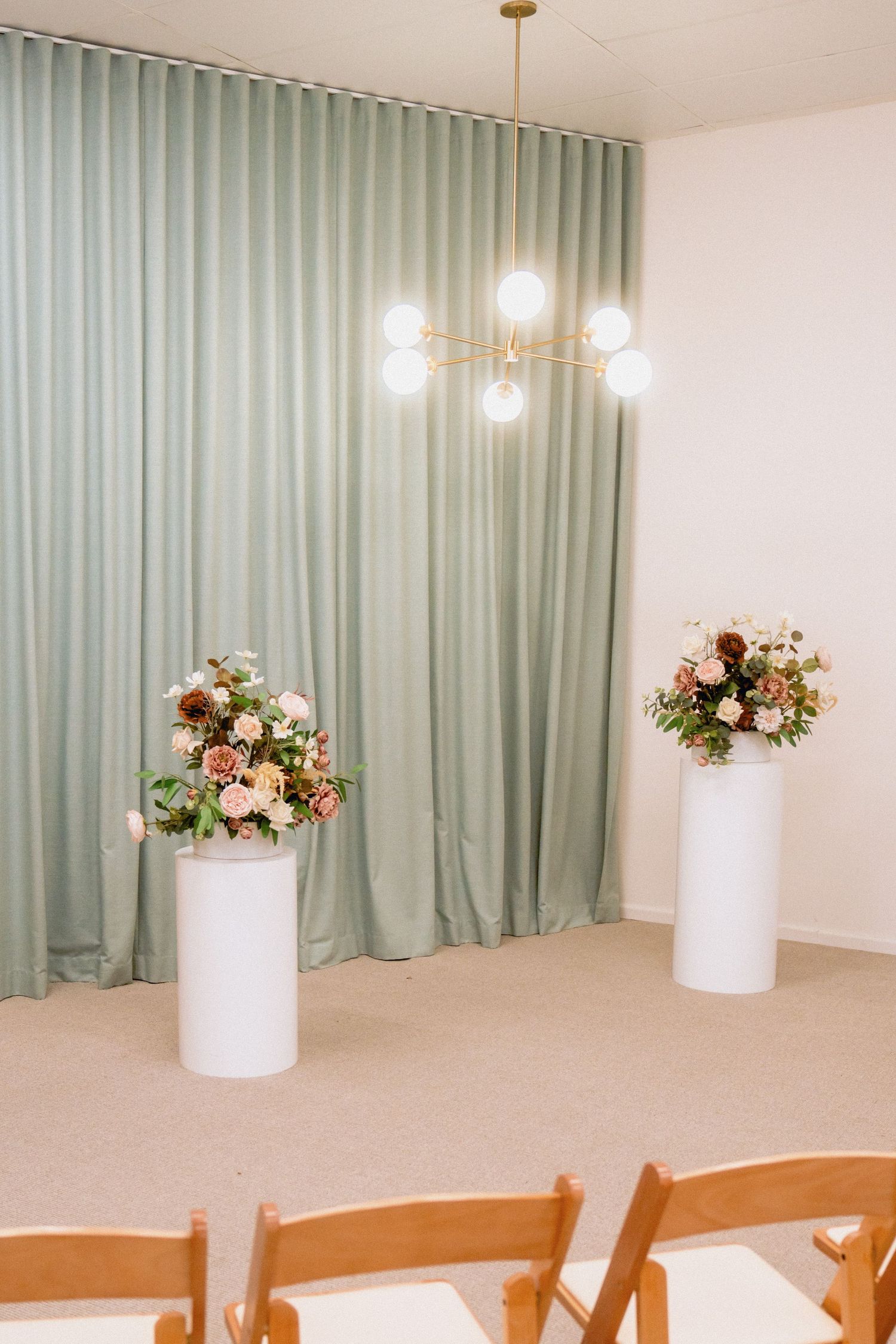 Elegant wedding venue setup with white floral arrangements on pedestals against sage green curtains.