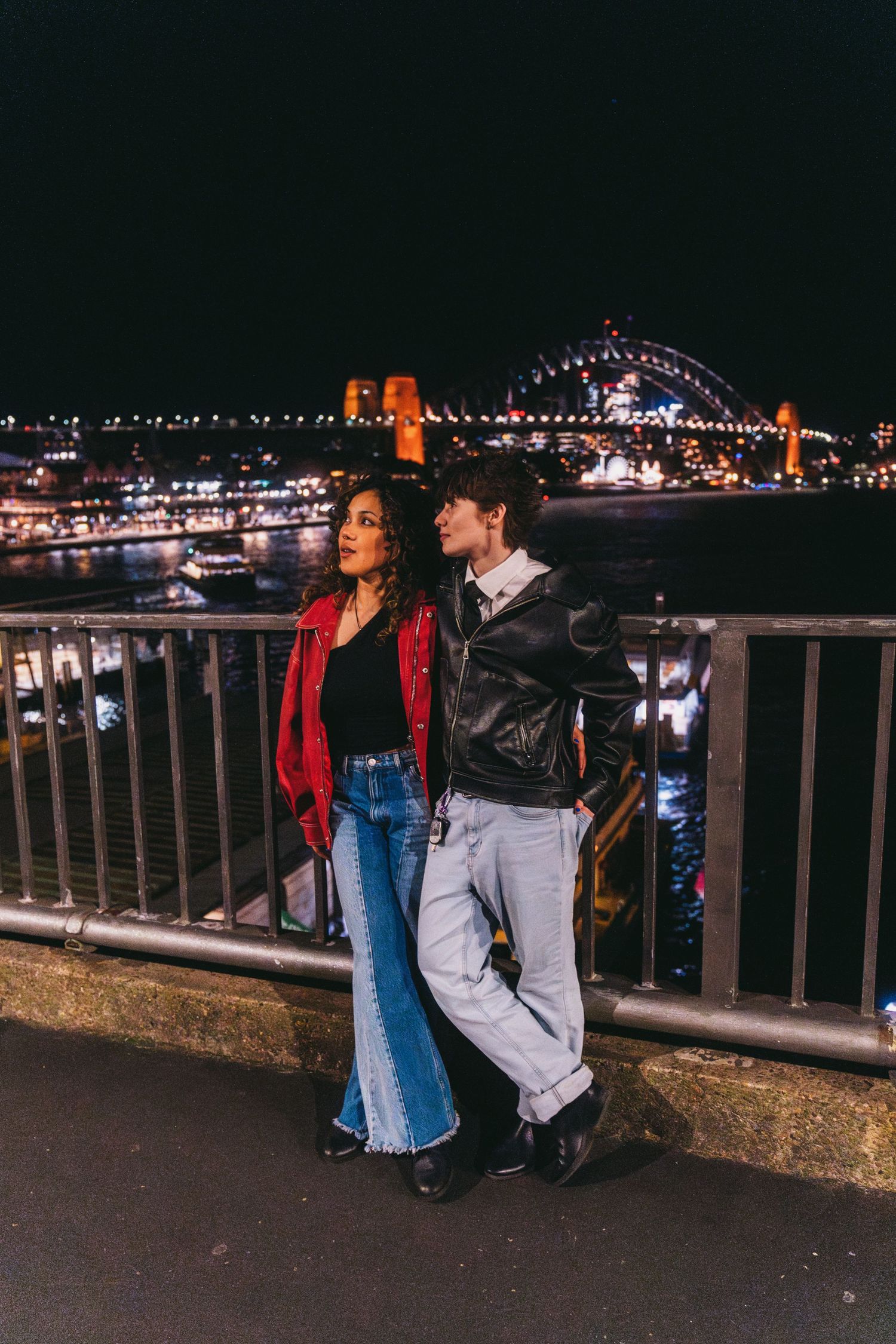 A couple stands by a railing with a illuminated bridge visible in the background.