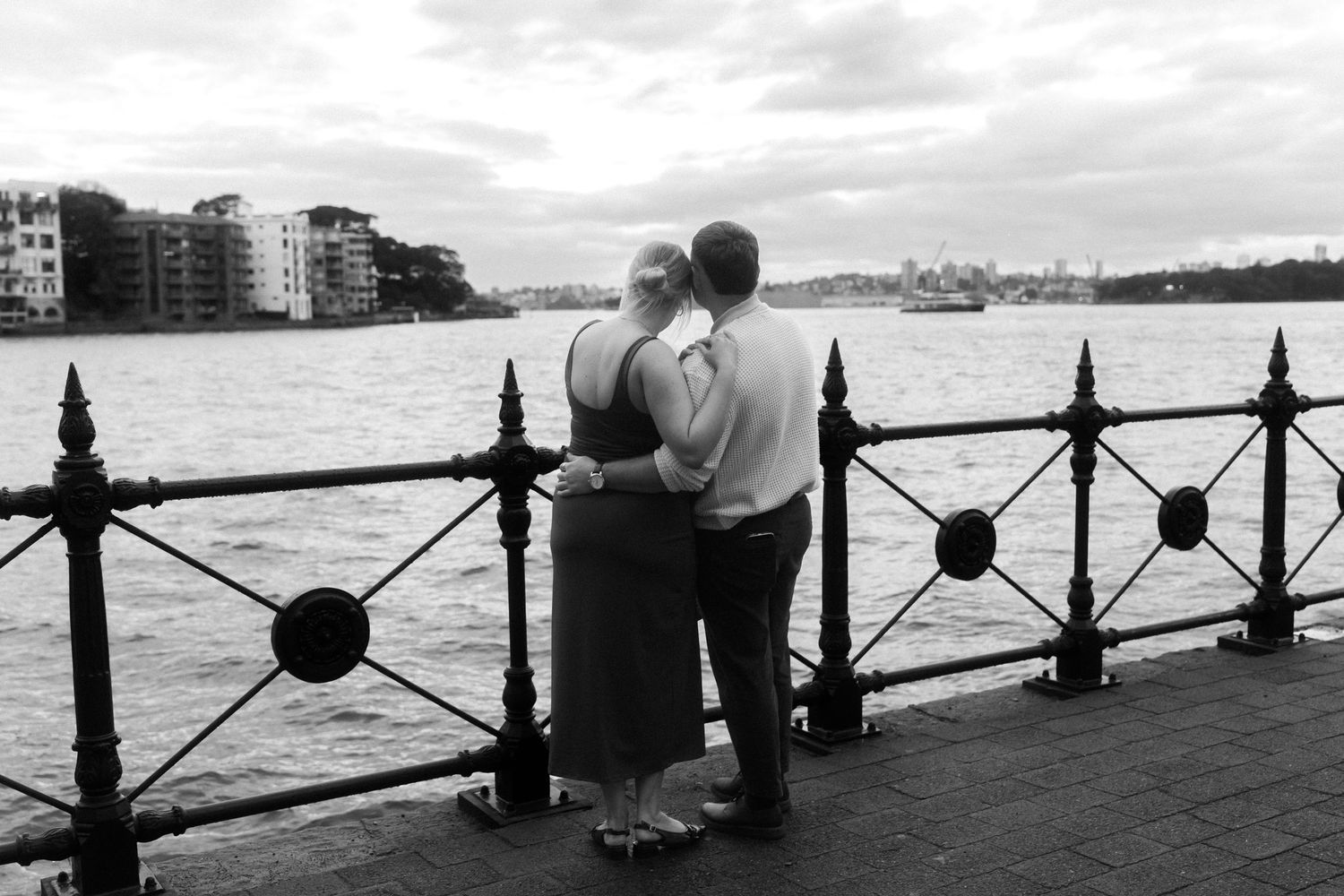 A panoramic black and white view of a couple by the waterfront with city buildings in background.