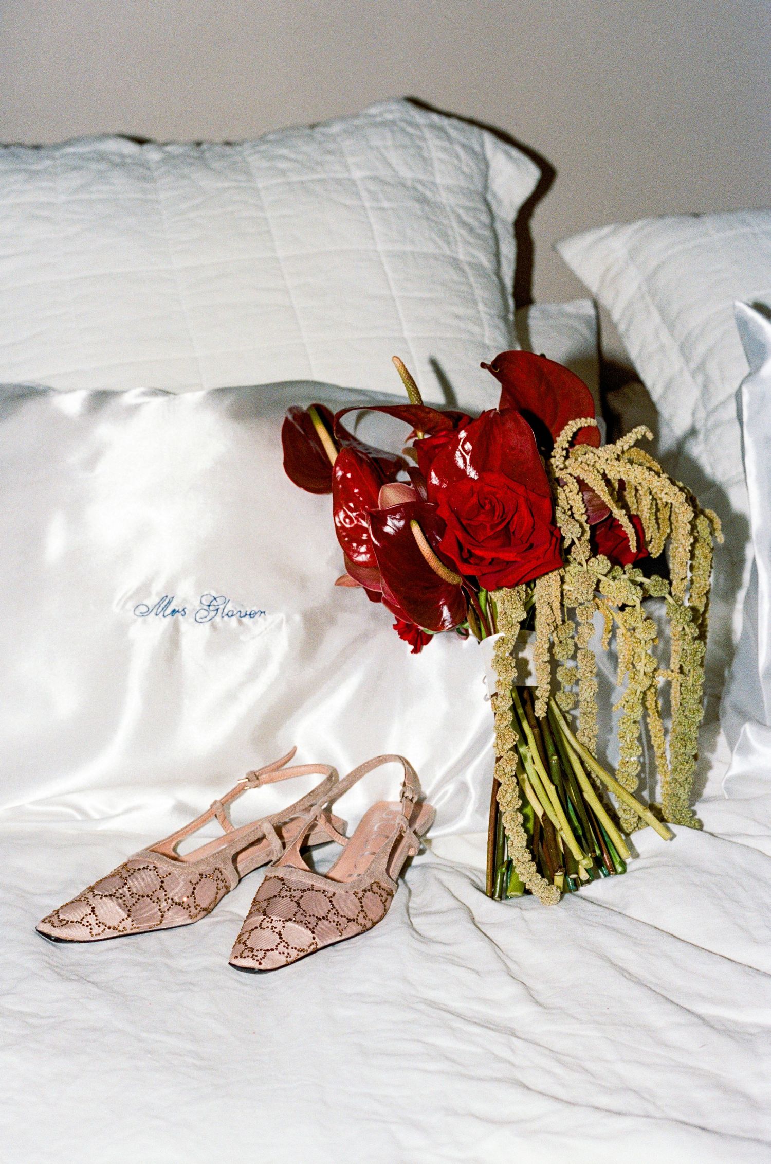 Wedding details with red roses, silk pillow, and pink snakeskin shoes on white bedding.