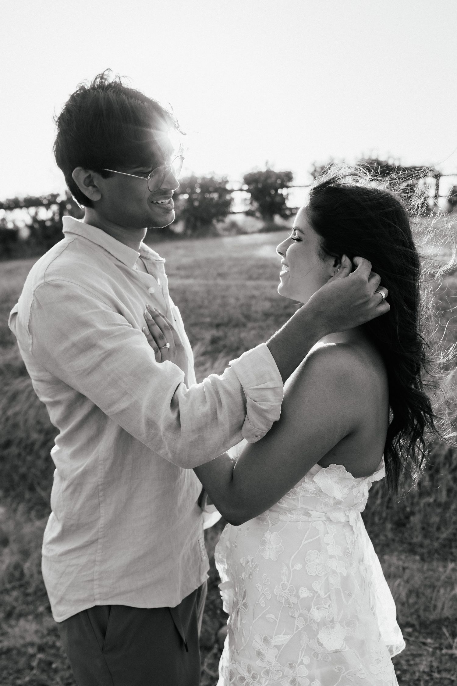 Playful couple in white clothing share an intimate moment in a field during a photo session.