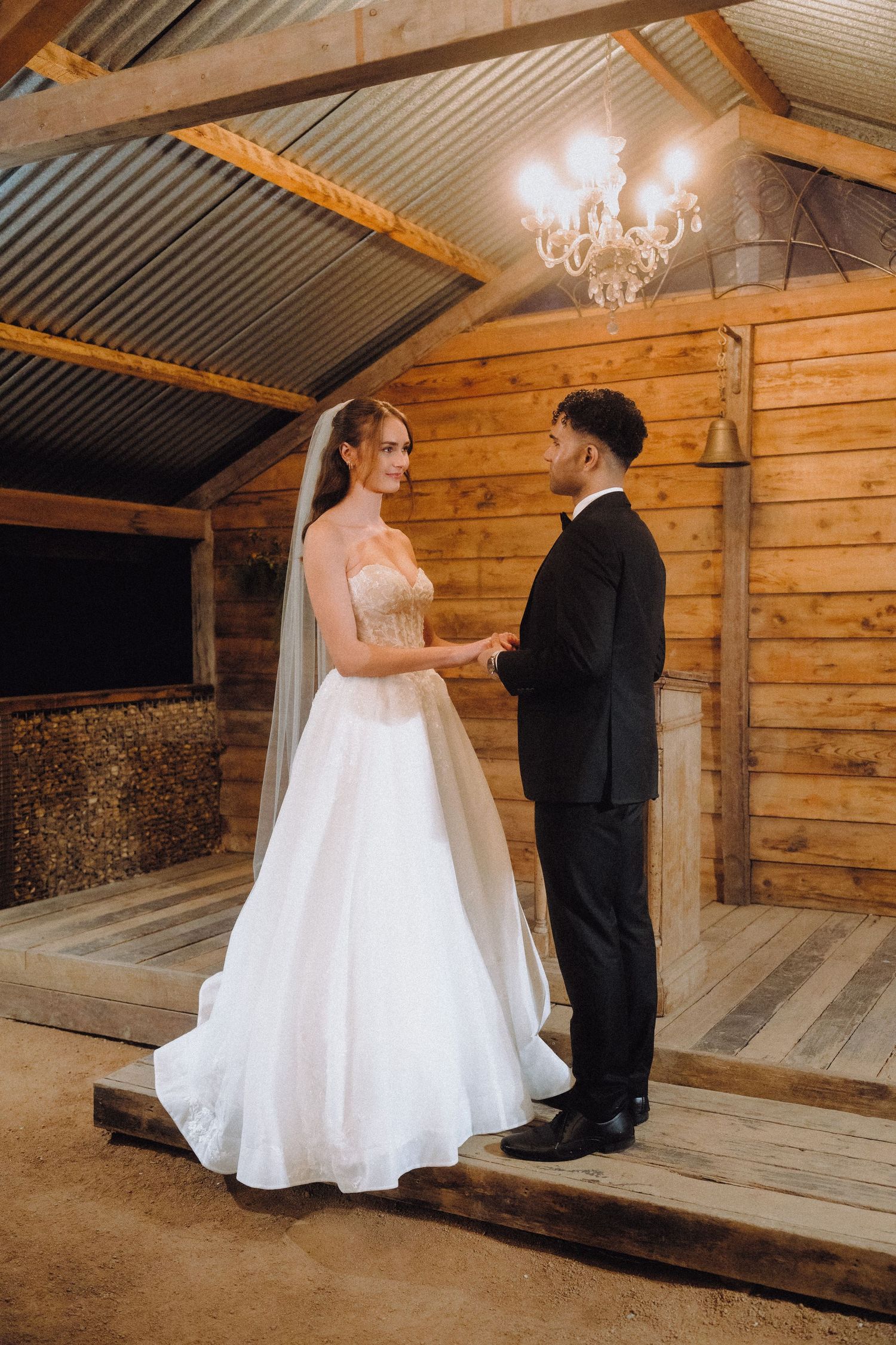 A couple shares a romantic moment in a rustic wooden barn setting with a chandelier overhead.