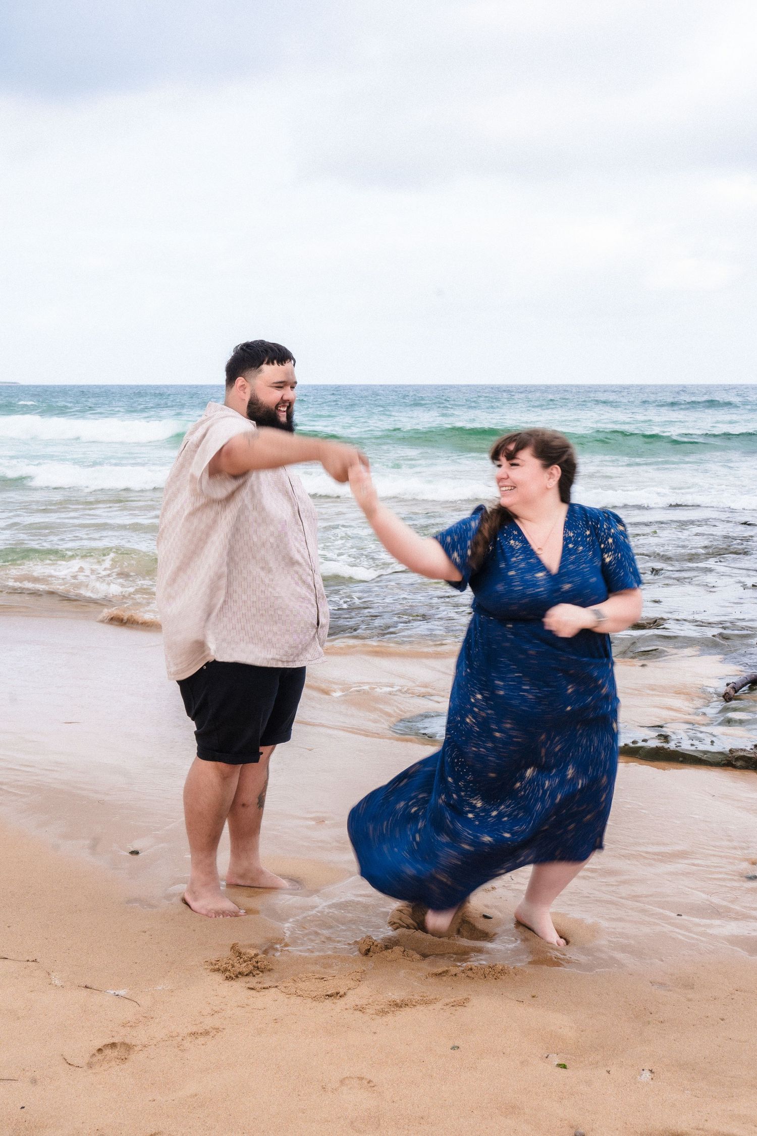 Playful couple splashing and dancing together on a sandy beach by the ocean waves.