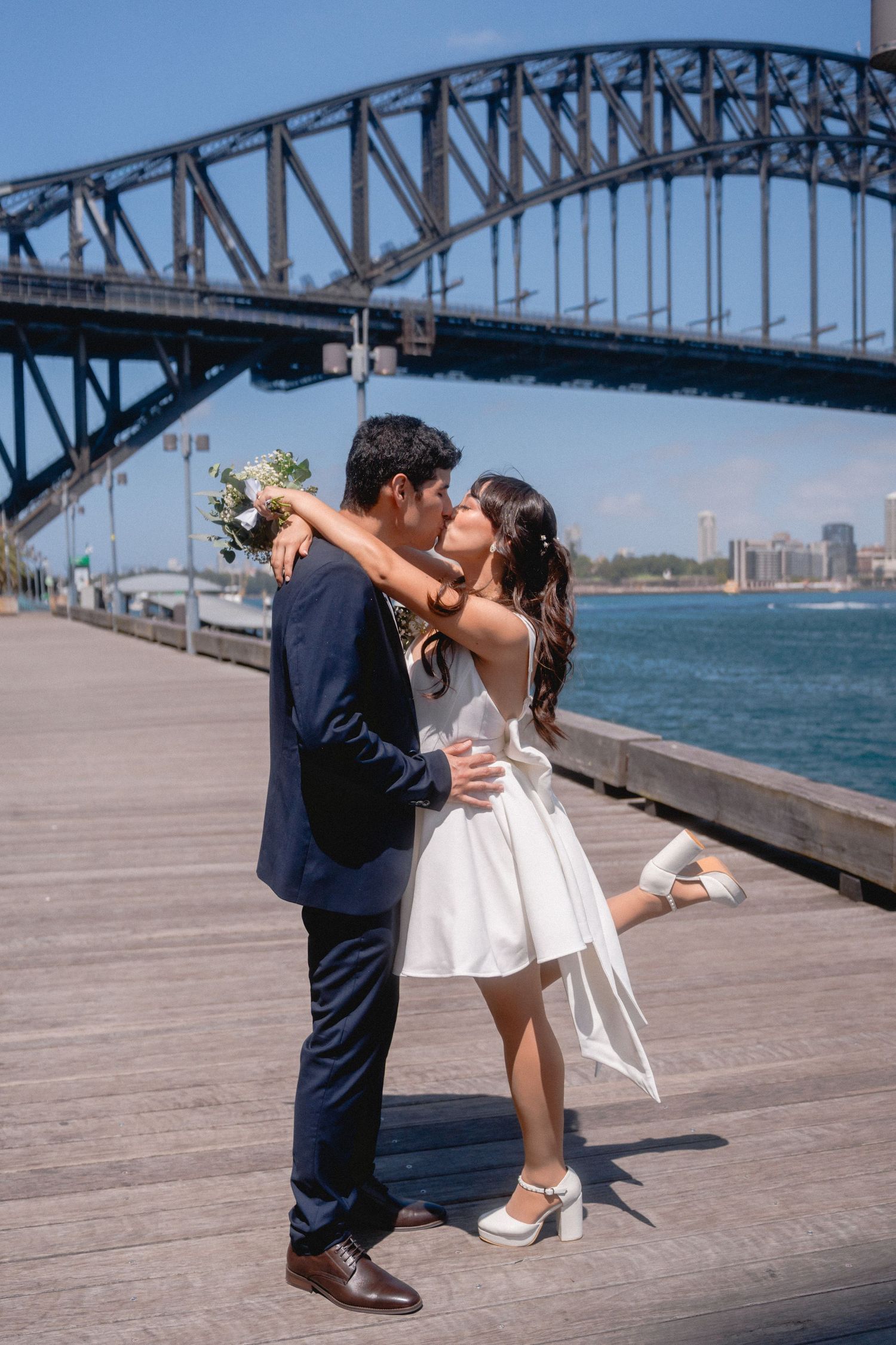 A couple shares romantic moments on a boardwalk with the Sydney Harbour Bridge in the background.