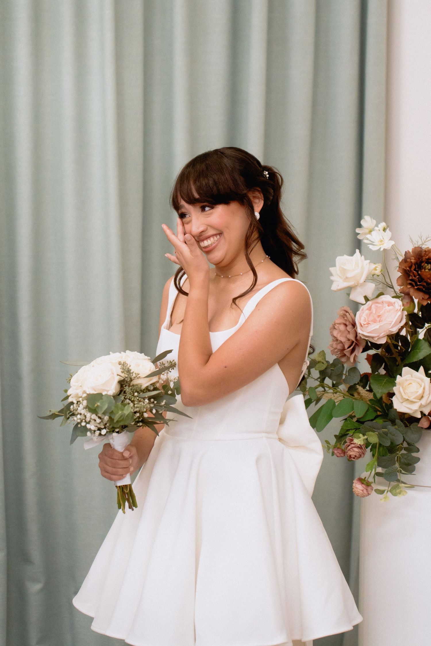 Joyful bride in white dress holding bouquet against sage green curtains.