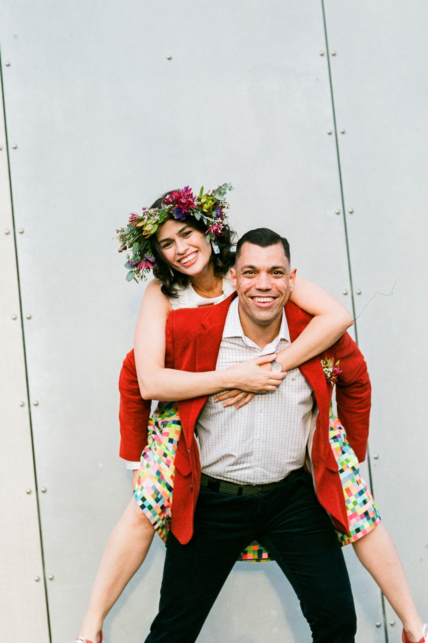 Playful wedding portrait with bright red jacket and floral crown against white wall.
