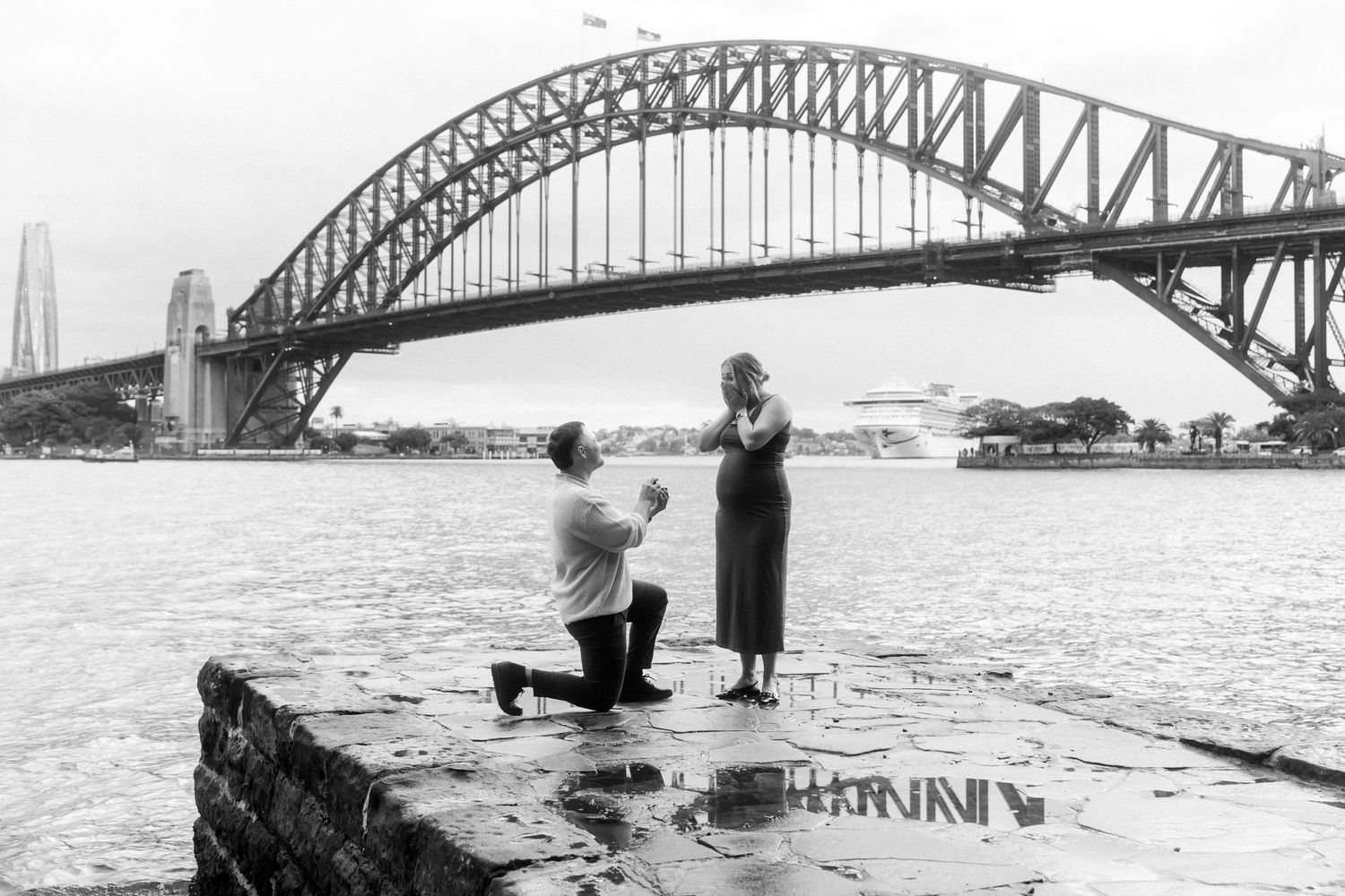 A black and white photo of a proposal moment in front of the Sydney Harbour Bridge.
