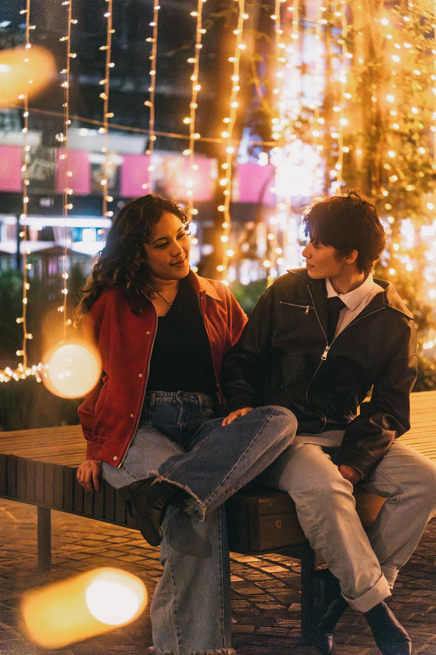 Two people share intimate conversation on bench surrounded by warm fairy lights.