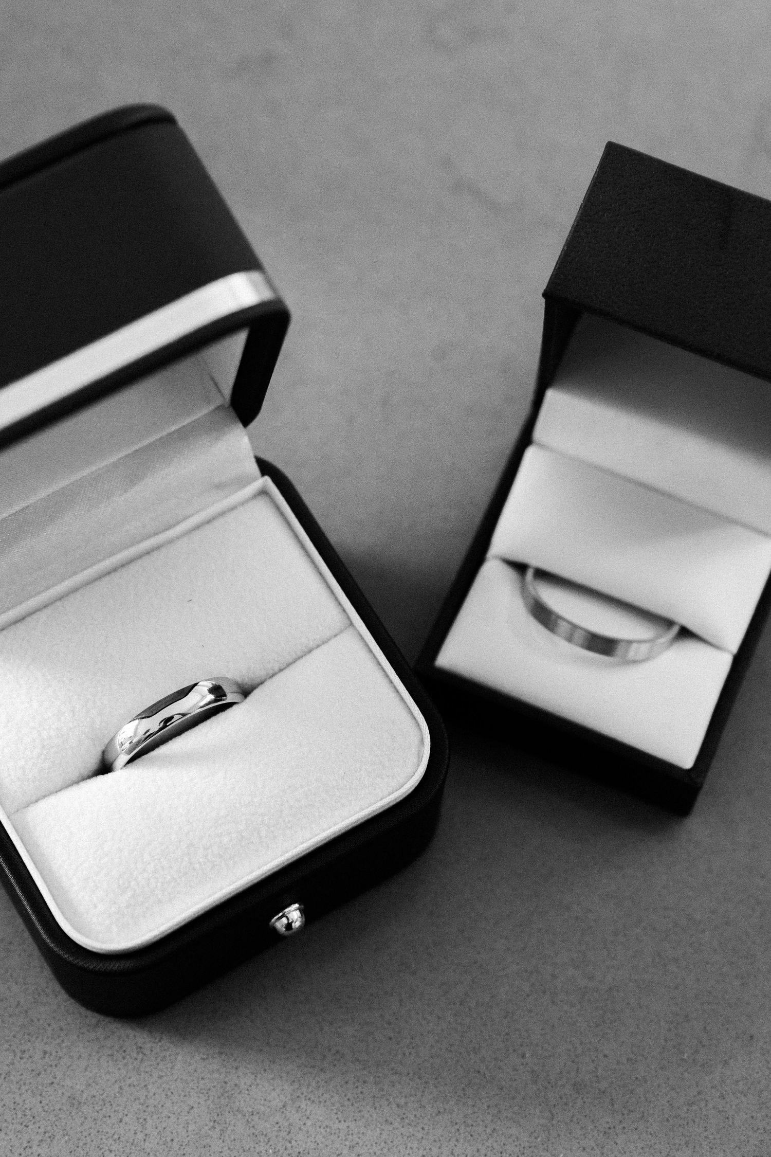 Two silver wedding bands displayed in open black ring boxes on a gray surface, black and white photo.