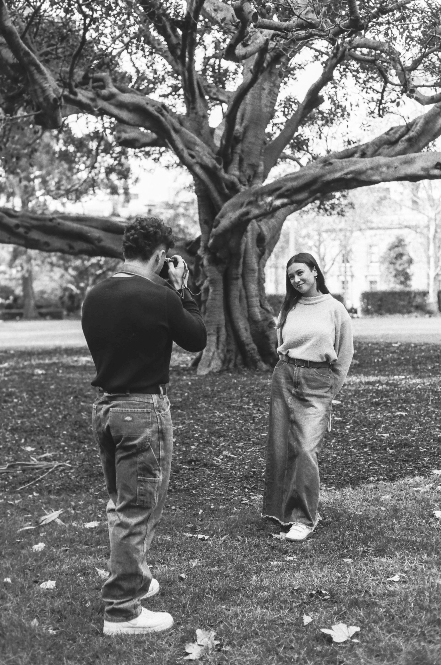 A photographer takes pictures of a model posing near a large tree in a park setting.