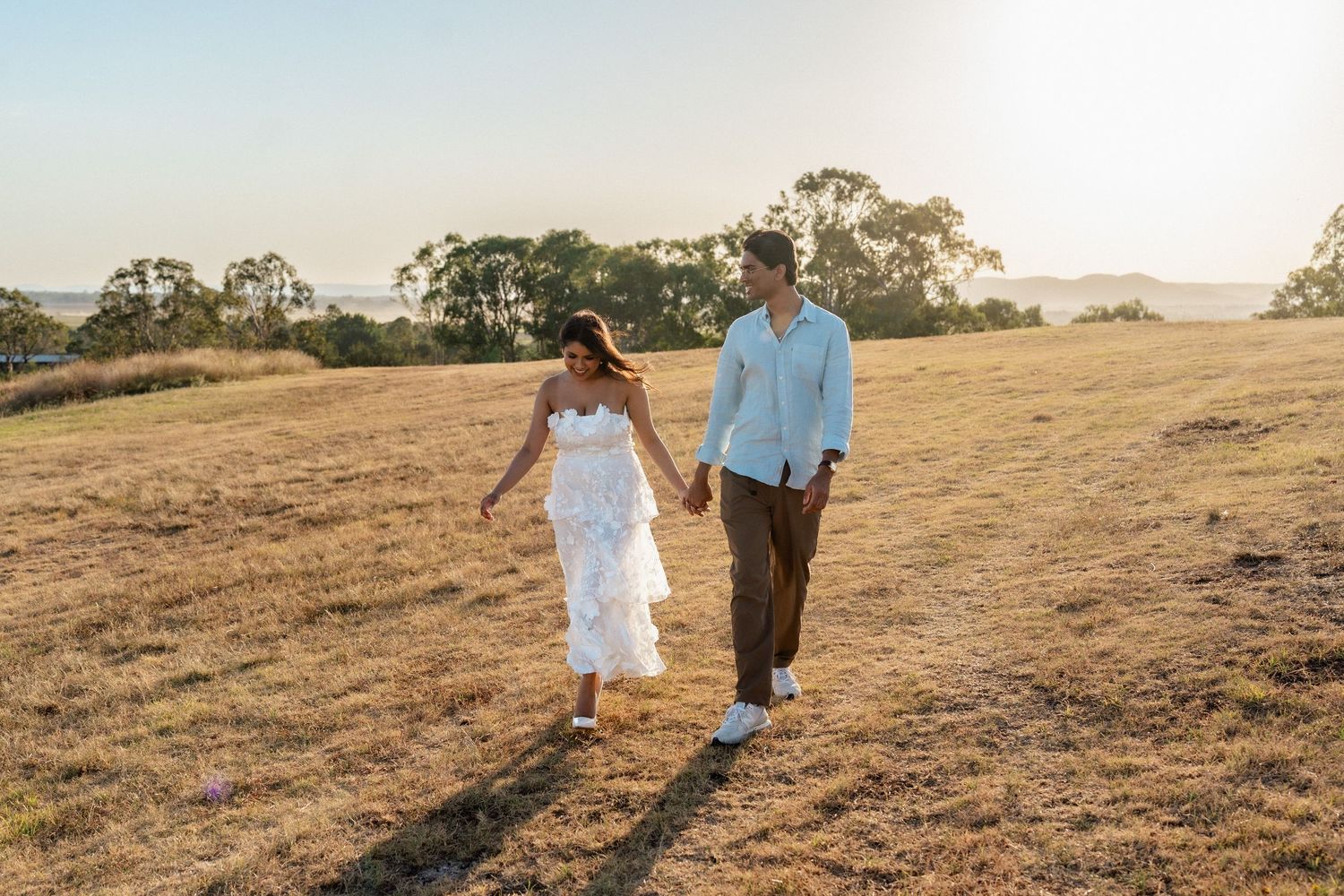 A couple walks hand in hand through a golden field at sunset creating long shadows on the ground.