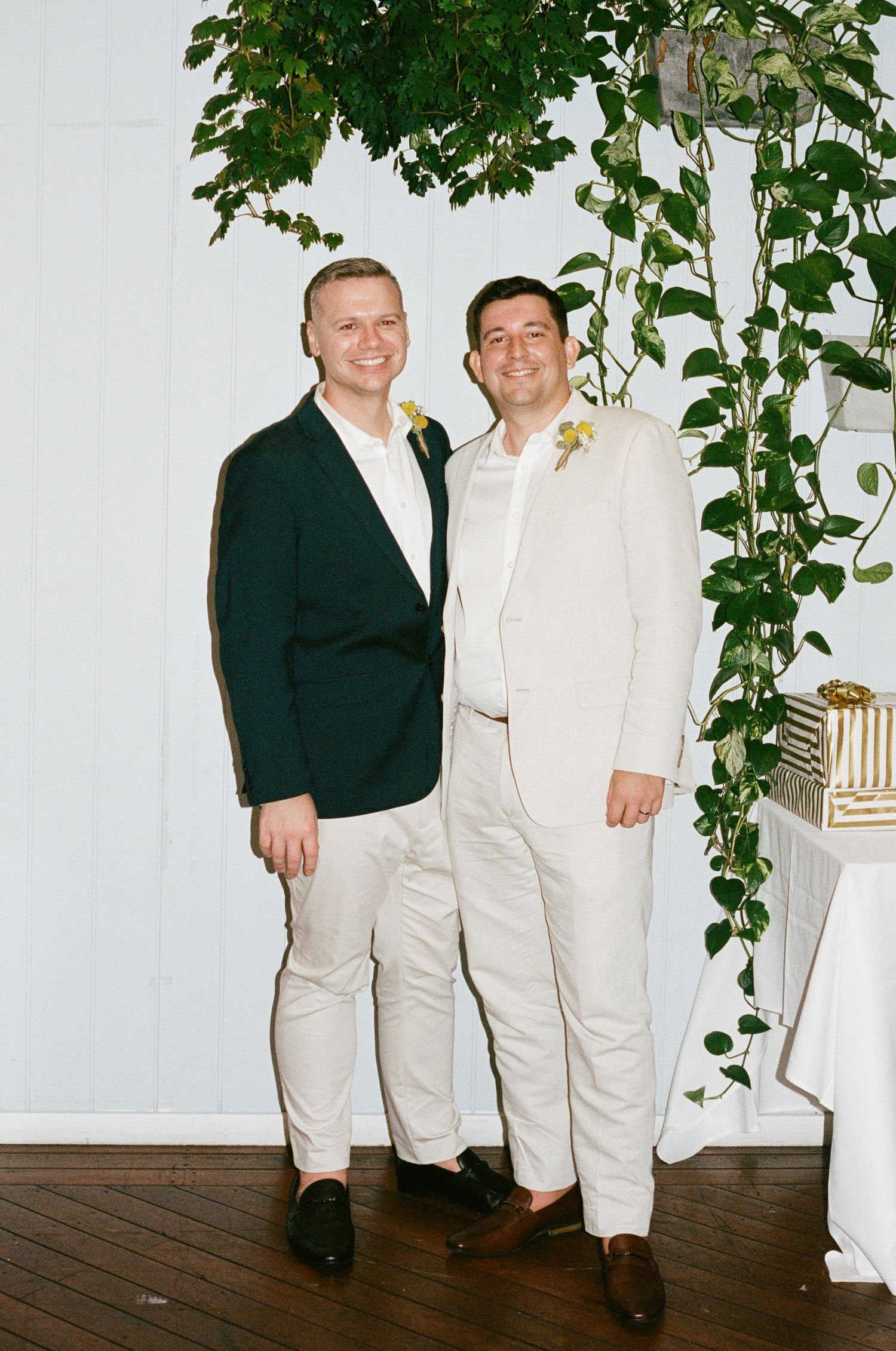 Two grooms smiling together at wedding ceremony, one in dark blazer, one in cream suit with boutonniere, surrounded by greenery.