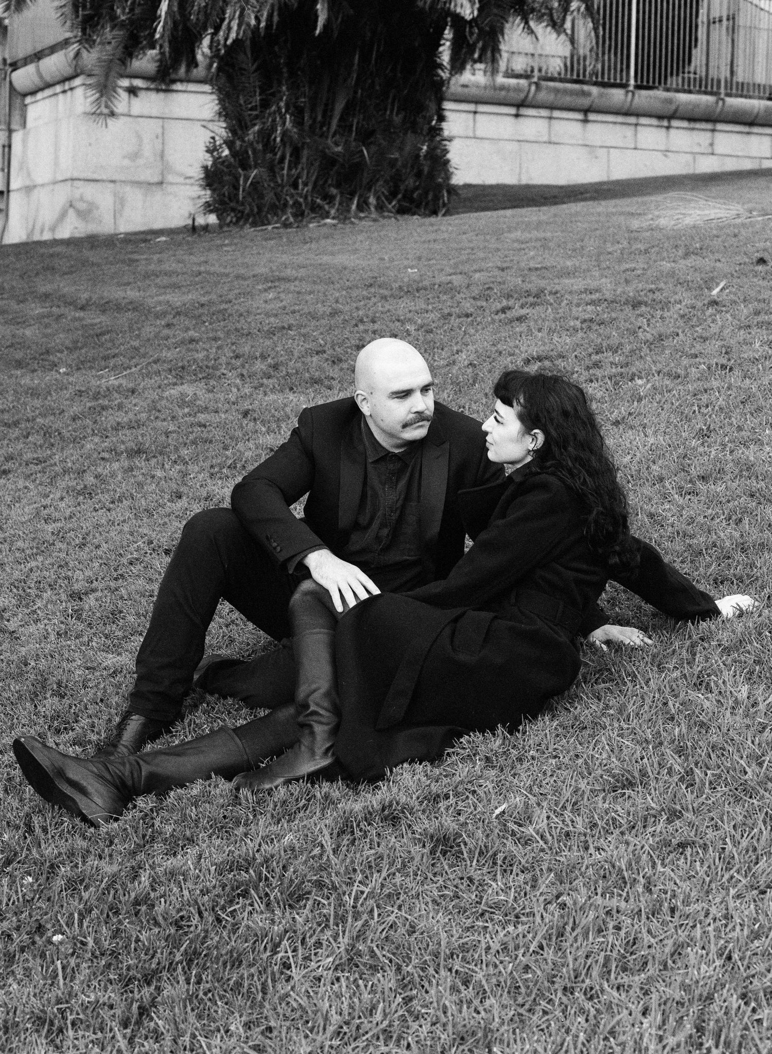 A couple in black clothing lies together on grass in an artistic black and white photo.