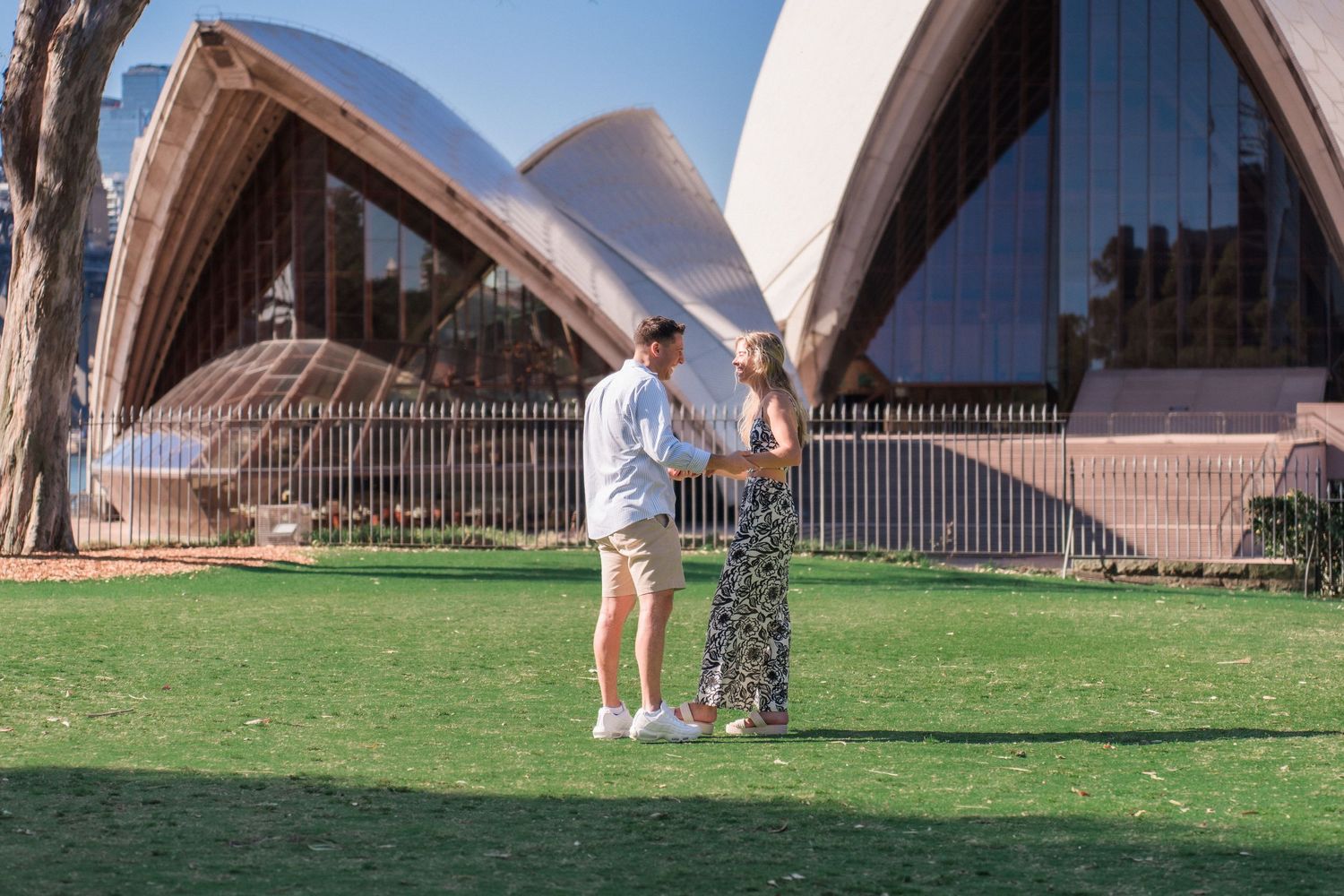 A romantic moment unfolds on grass with Sydney Opera House visible in background.