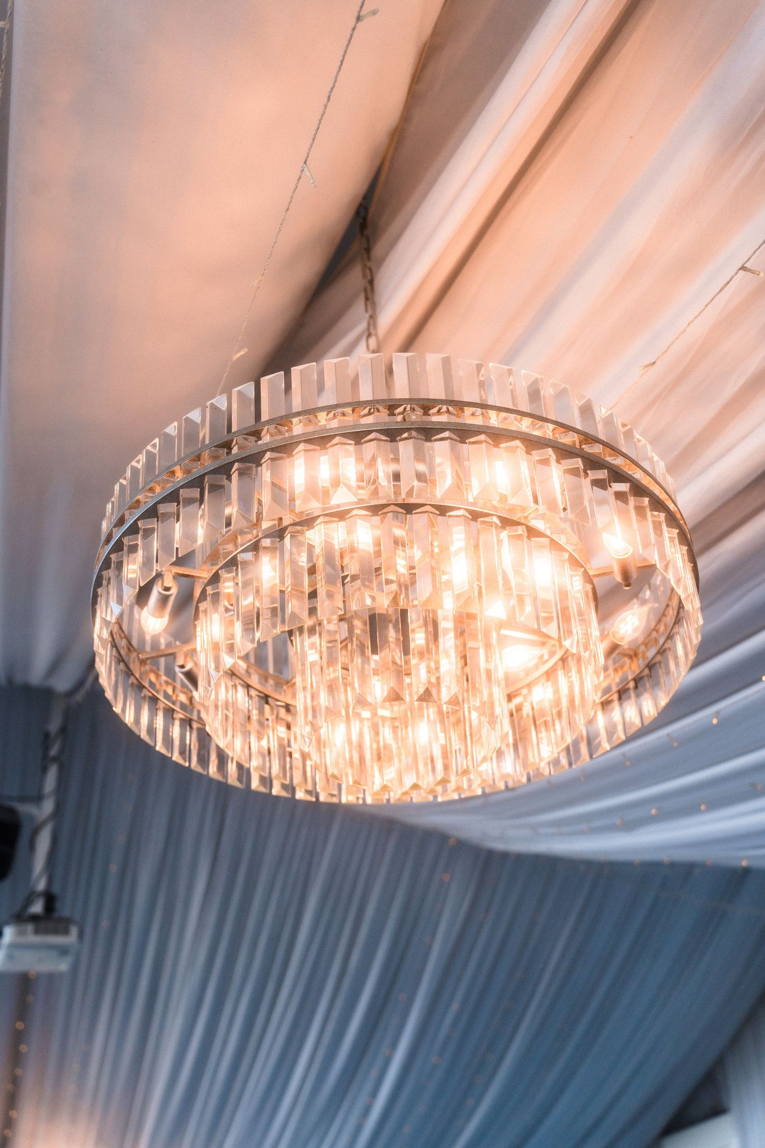 Crystal chandelier with tiered glass prisms hanging from draped white ceiling in elegant event space.