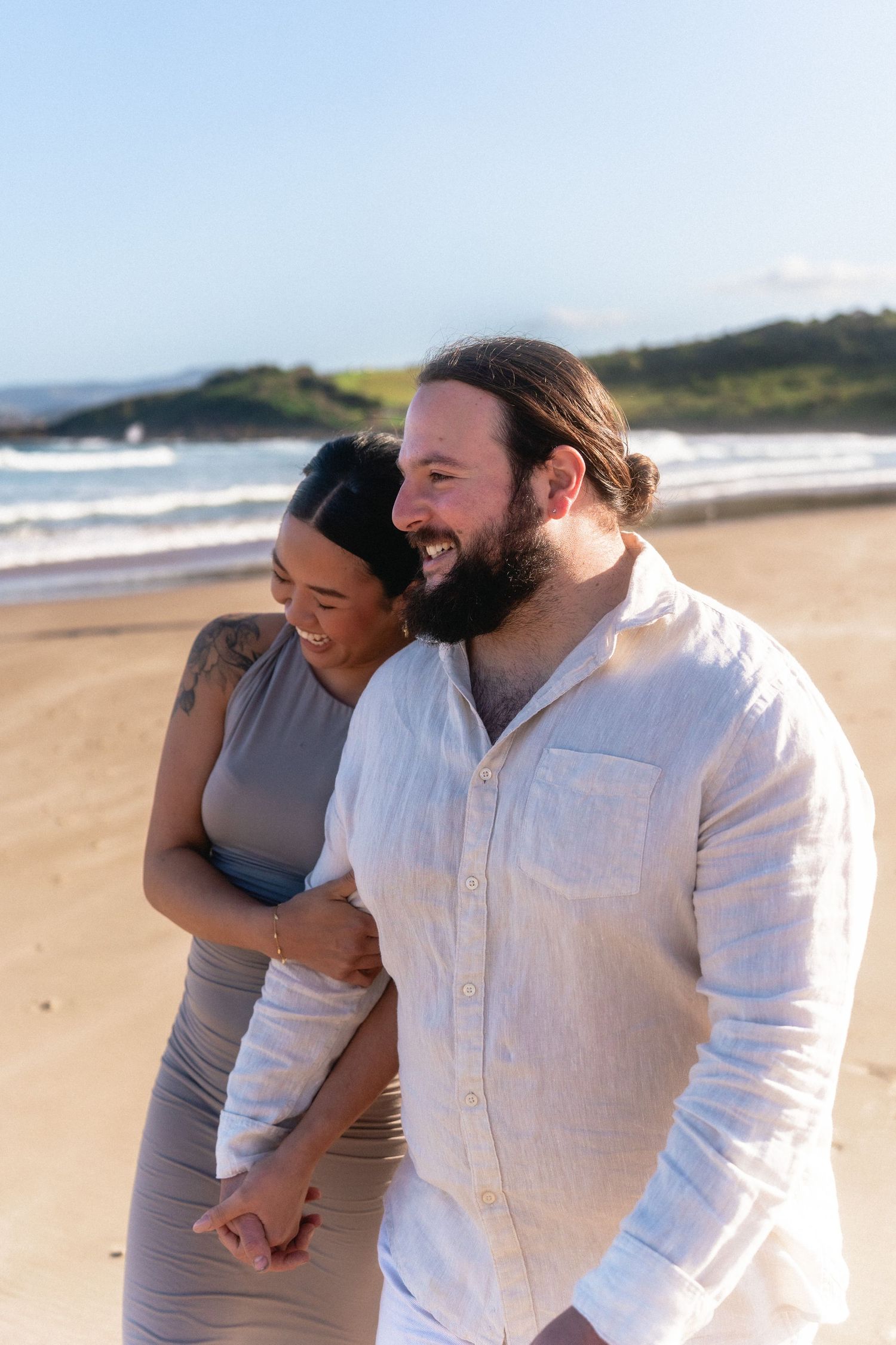 A joyful couple laughs together on a sunny beach with ocean waves in the background.