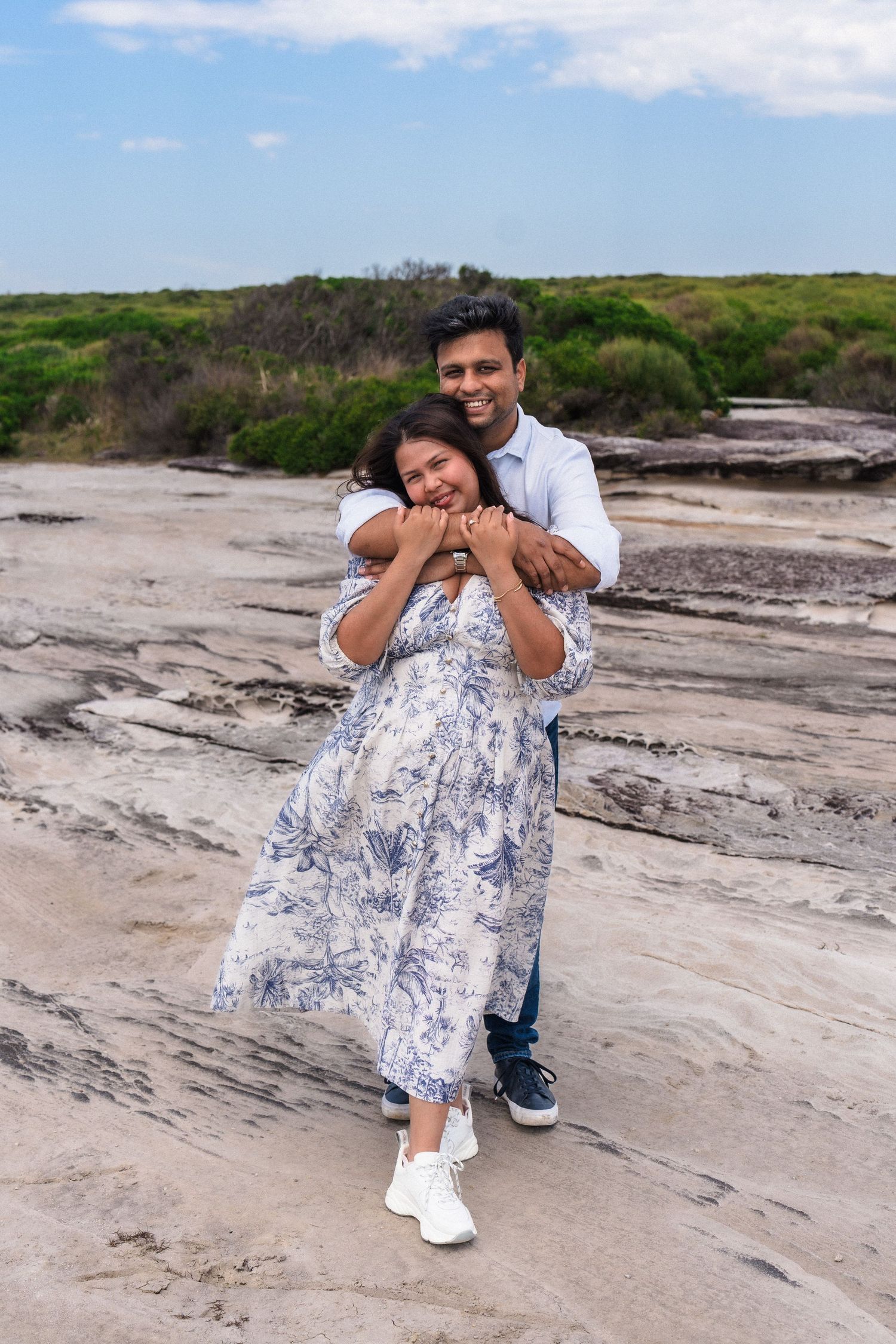 A couple poses playfully on rocky coastal terrain wearing blue and white outfits.