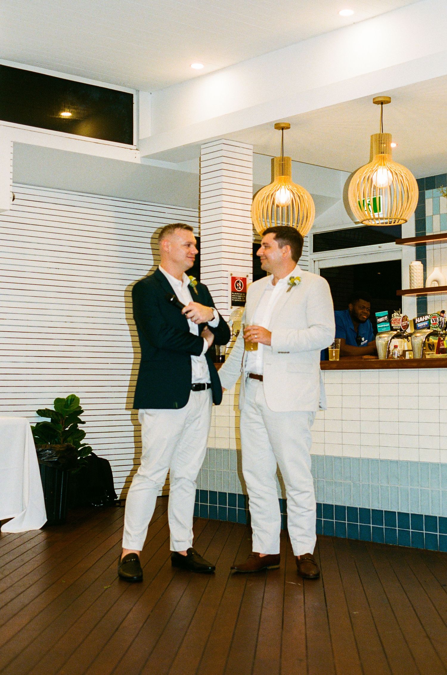 Two grooms in white and navy suits laughing together at their wedding reception near a stylish bar.