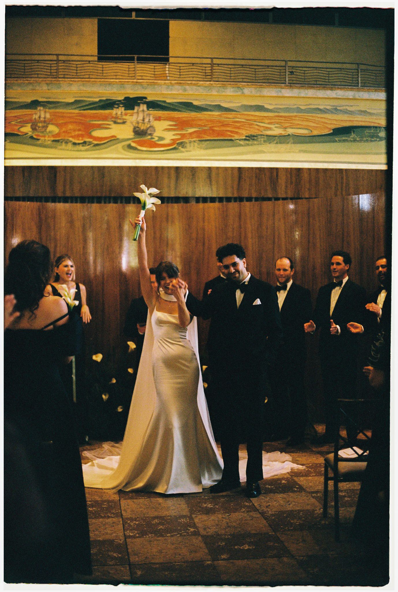Bride raises bouquet triumphantly as couple walks down aisle after wedding ceremony in elegant wood-paneled venue.