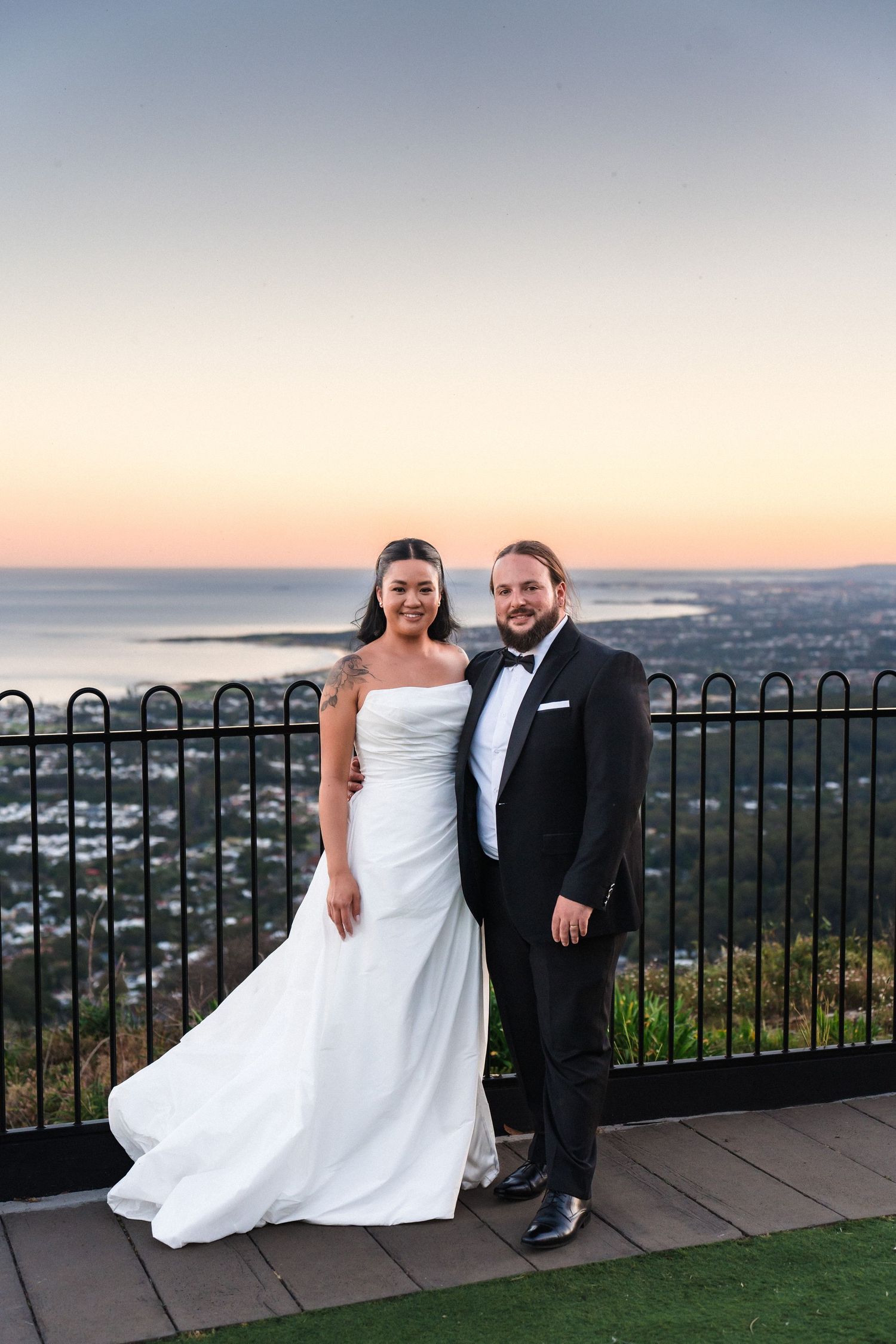 Elegant couple in formal wedding attire poses against scenic coastal sunset backdrop.