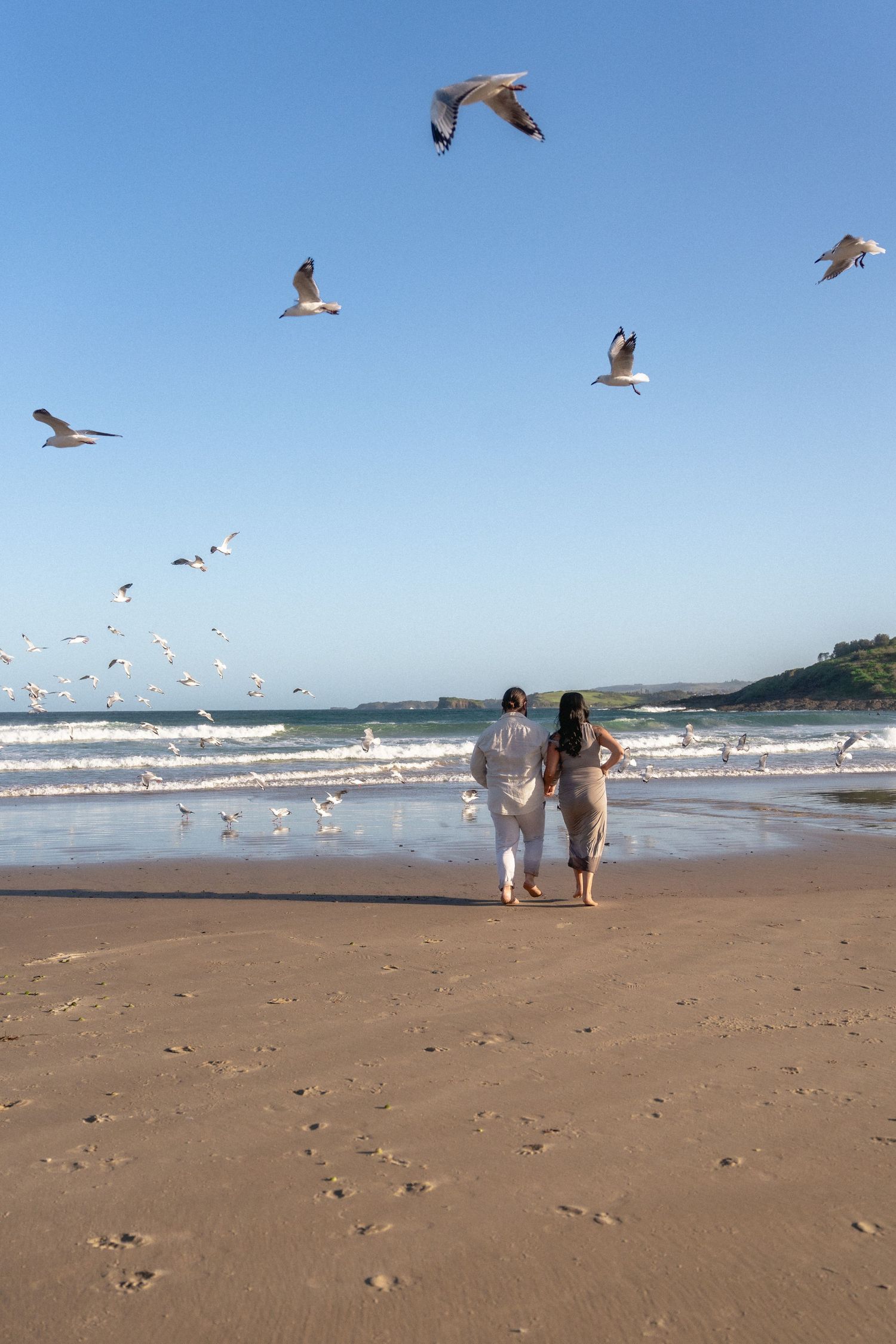 A couple walks on a beach while seagulls fly overhead.